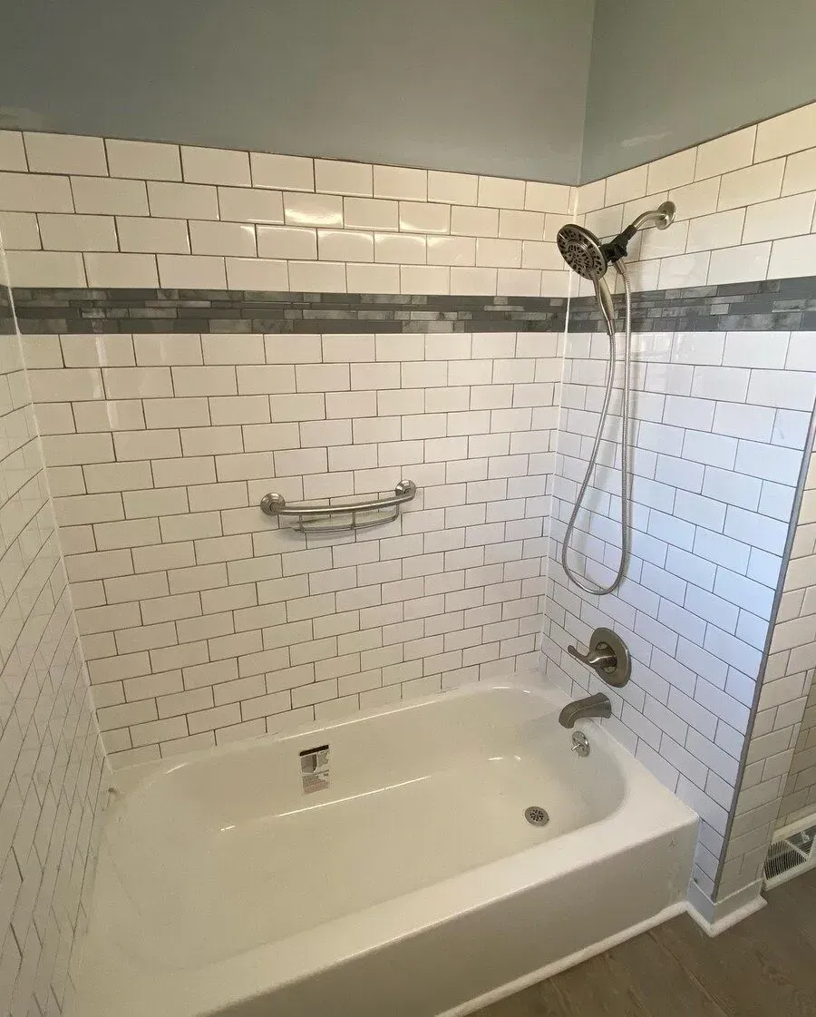 White tiled bathroom with a bathtub, showerhead, and chrome fixtures under a gray accent band