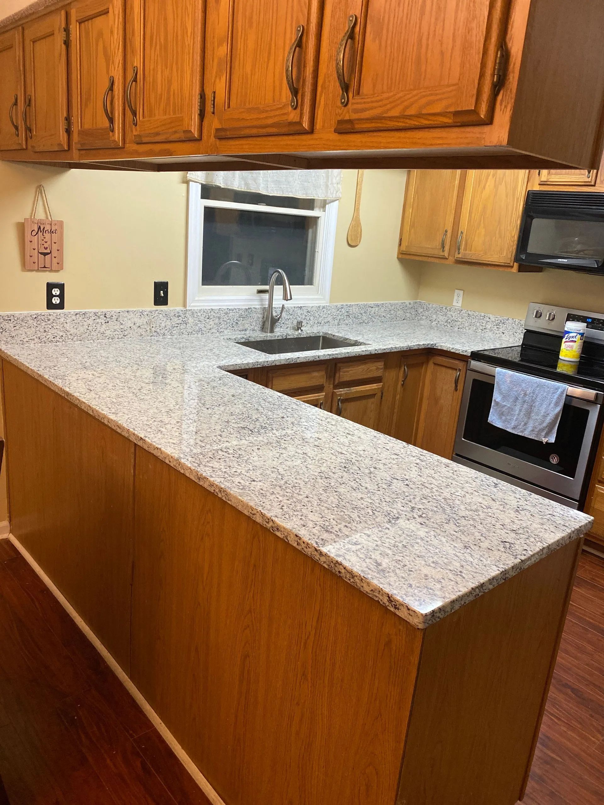 Kitchen with speckled granite countertops, wood cabinets, sink under window, and stove with oven mitts attached.