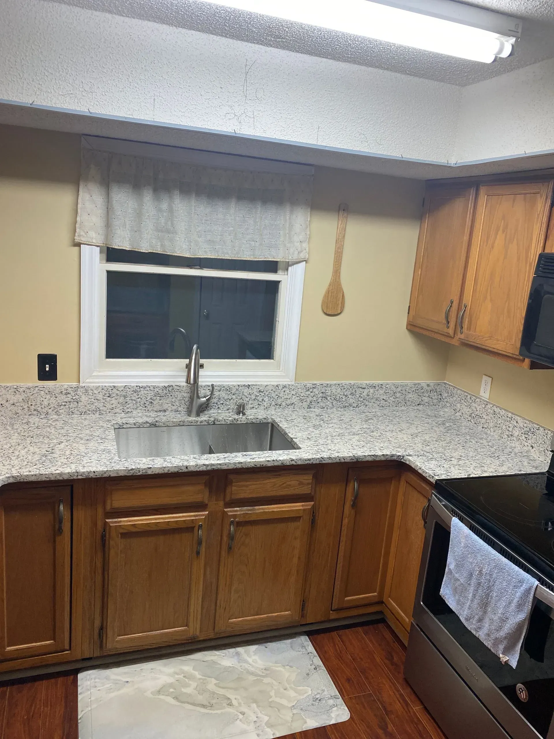 Small kitchen with wood cabinets, speckled countertops, sink under window, and stove on the right.
