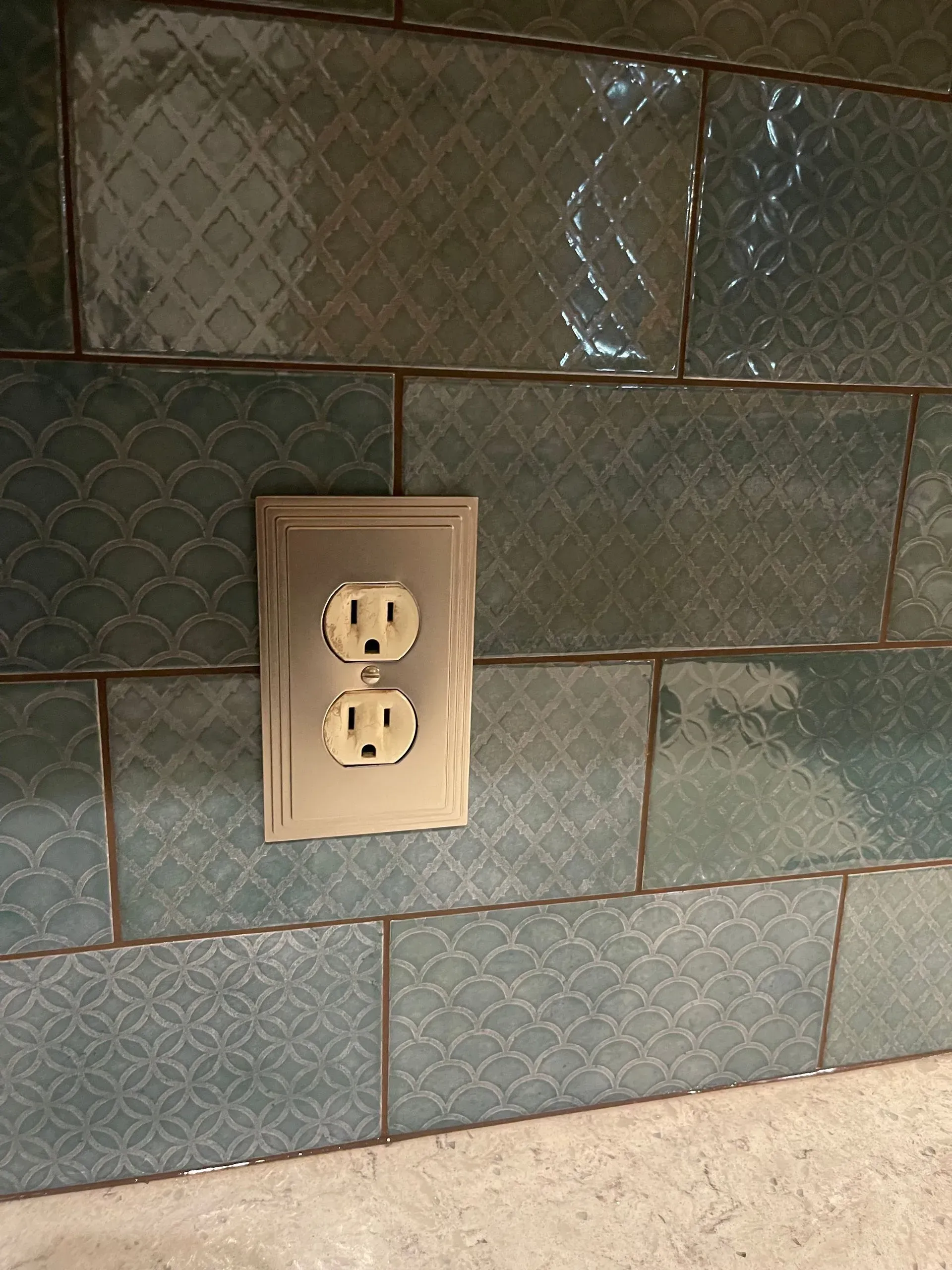 Beige wall outlet on blue-green tiled kitchen backsplash above a countertop