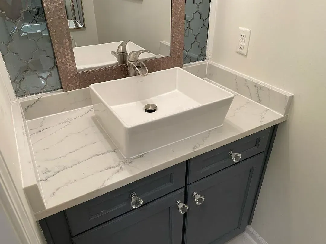 Bathroom vanity with white sink, marble countertop, and dark blue cabinets beside a mirror.