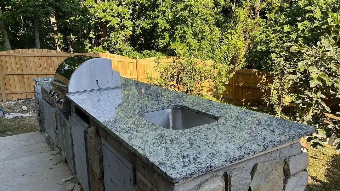 Outdoor stone counter with a built-in grill and sink, set in a backyard by a wooden fence.