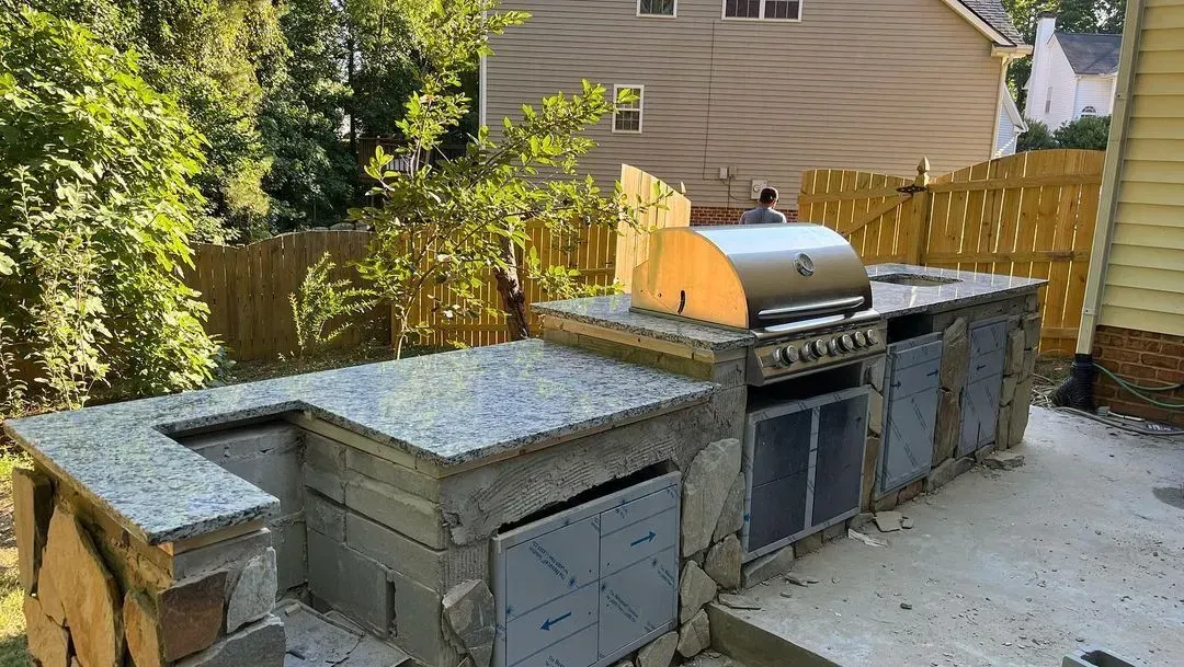 Outdoor backyard kitchen with stone counters, built-in grill, and a concrete patio beside a wooden fence.