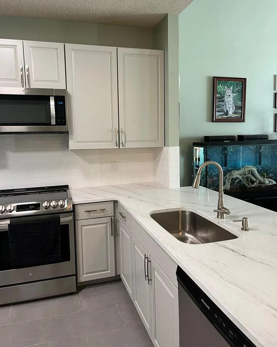 Modern kitchen with white cabinets, stainless stove, marble countertop, and sink by an aquarium.