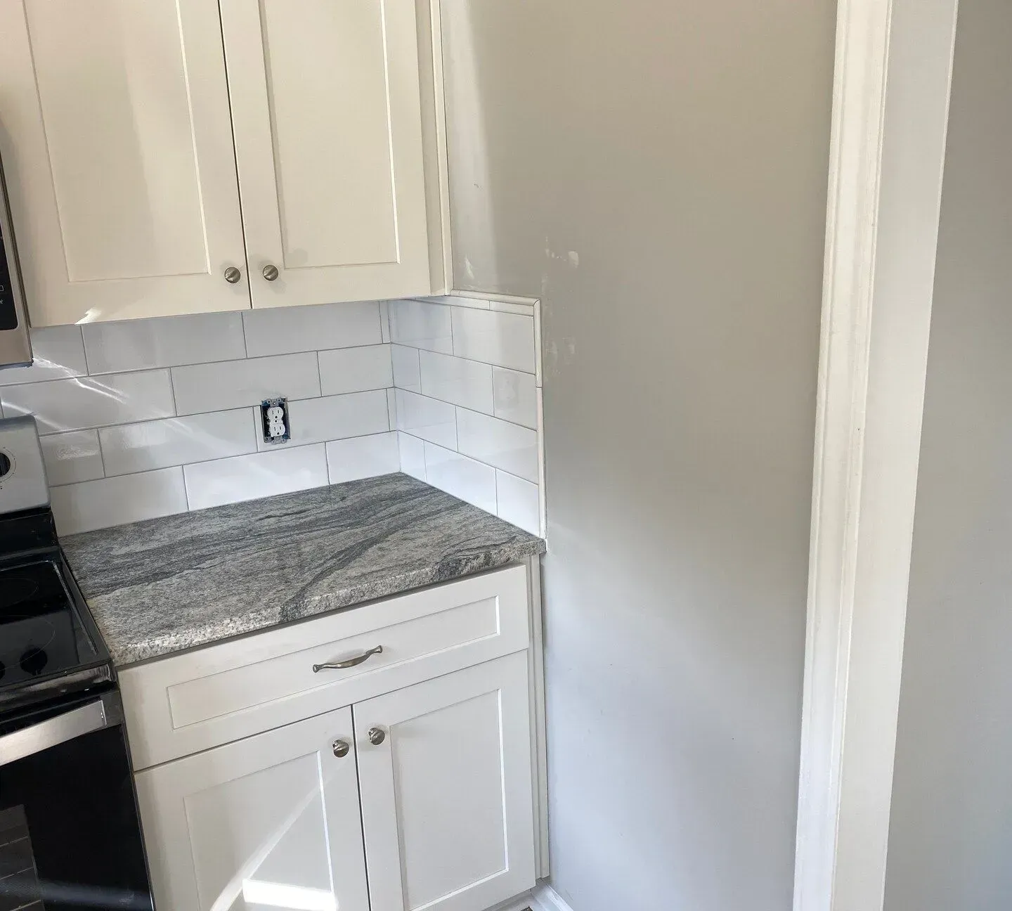 White kitchen corner with cabinets, granite countertop, and subway tile backsplash next to a gray wall