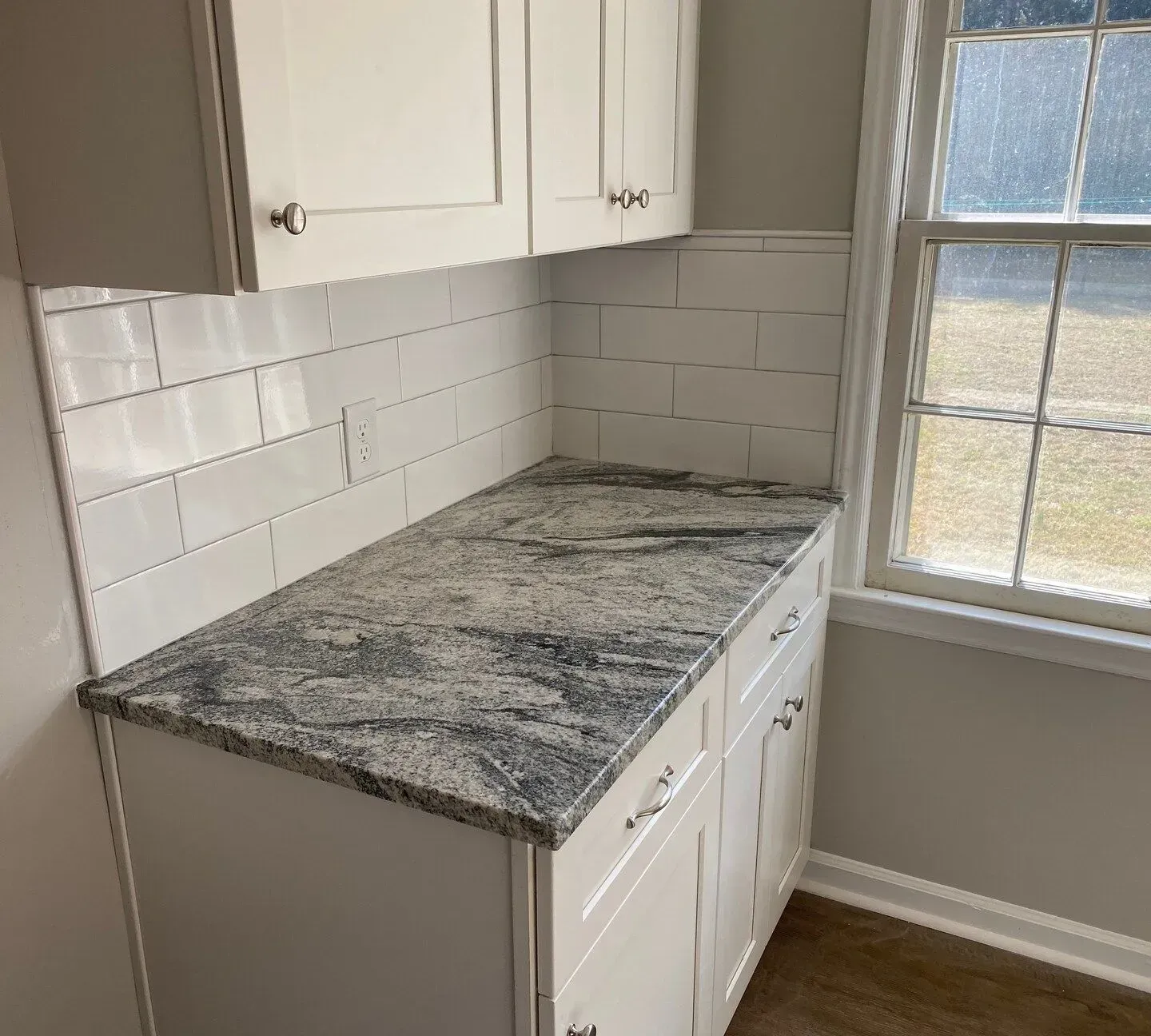 Small kitchen counter with granite top, white cabinets, subway tile backsplash, and window light