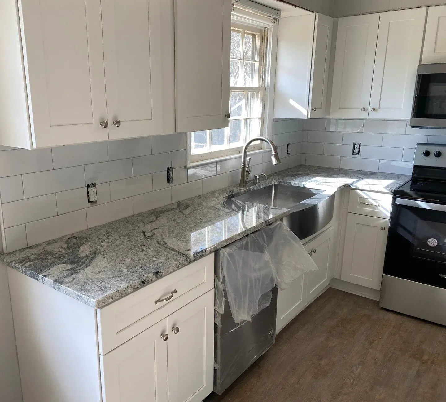 Bright white kitchen with granite countertops, stainless sink, dishwasher, and stove by a window.