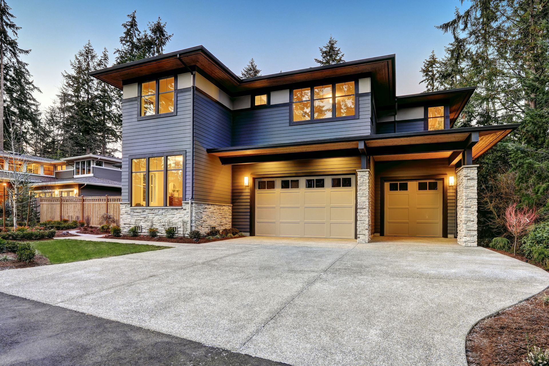 Modern two-story house with stone and blue siding, lit windows, and a three-car garage at dusk
