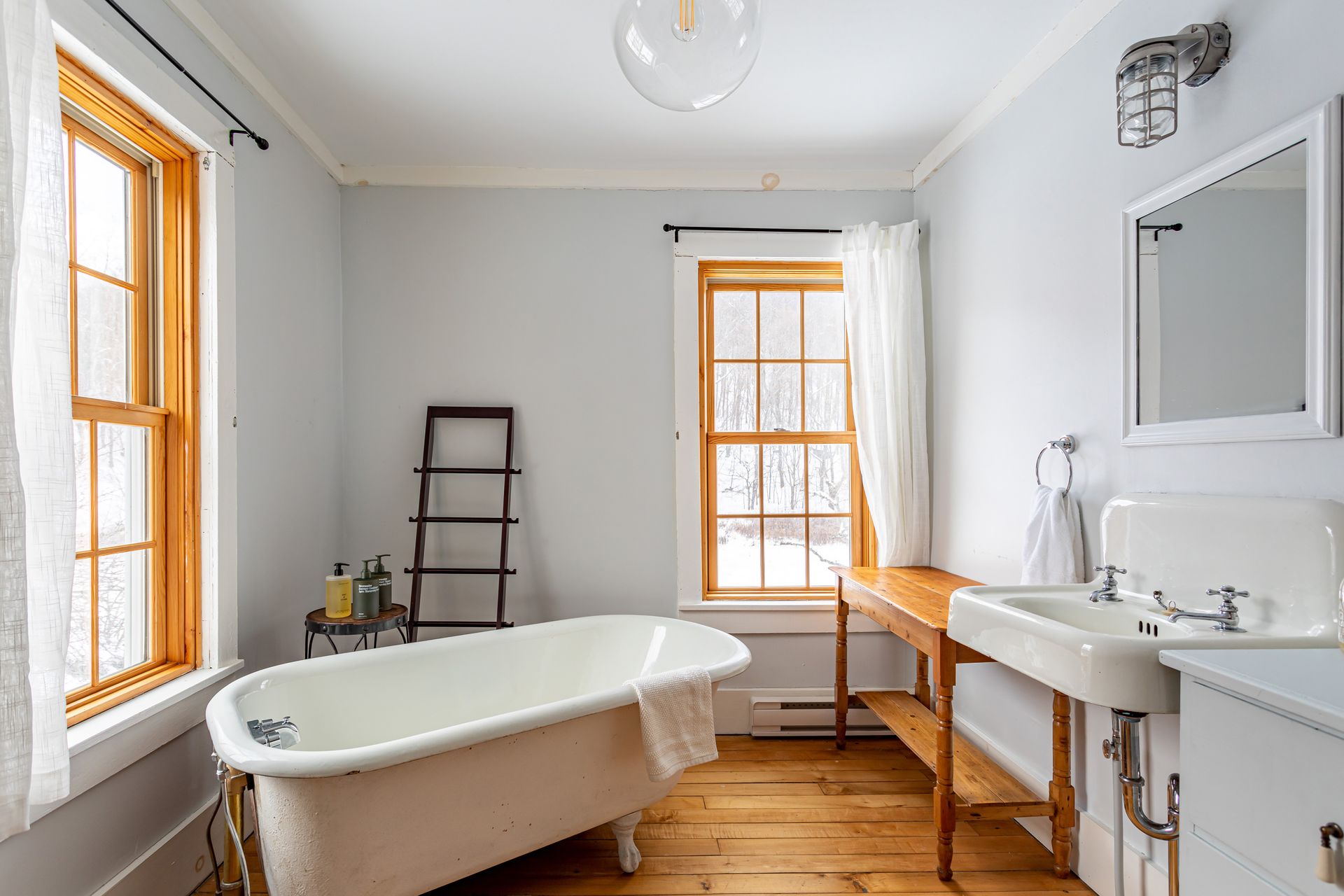 Bright bathroom with a clawfoot tub, white sink, wooden floor, and orange-trimmed windows