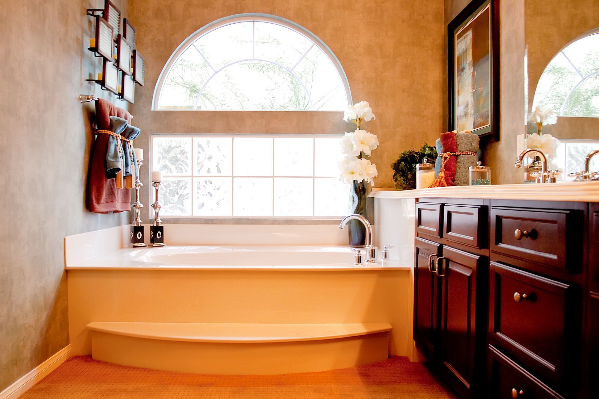 Bright bathroom with a white soaking tub beneath an arched window and dark wood cabinets on the right