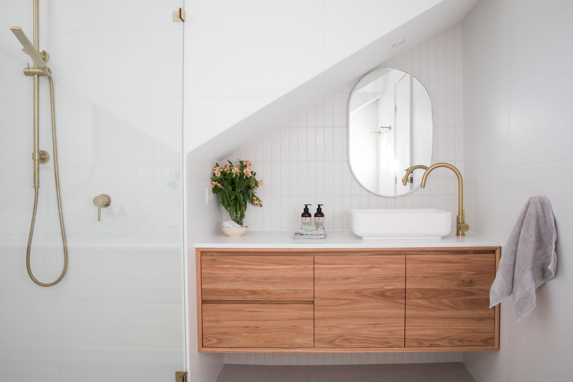 Modern white bathroom with wood vanity, round mirror, brass faucet, shower, and towel