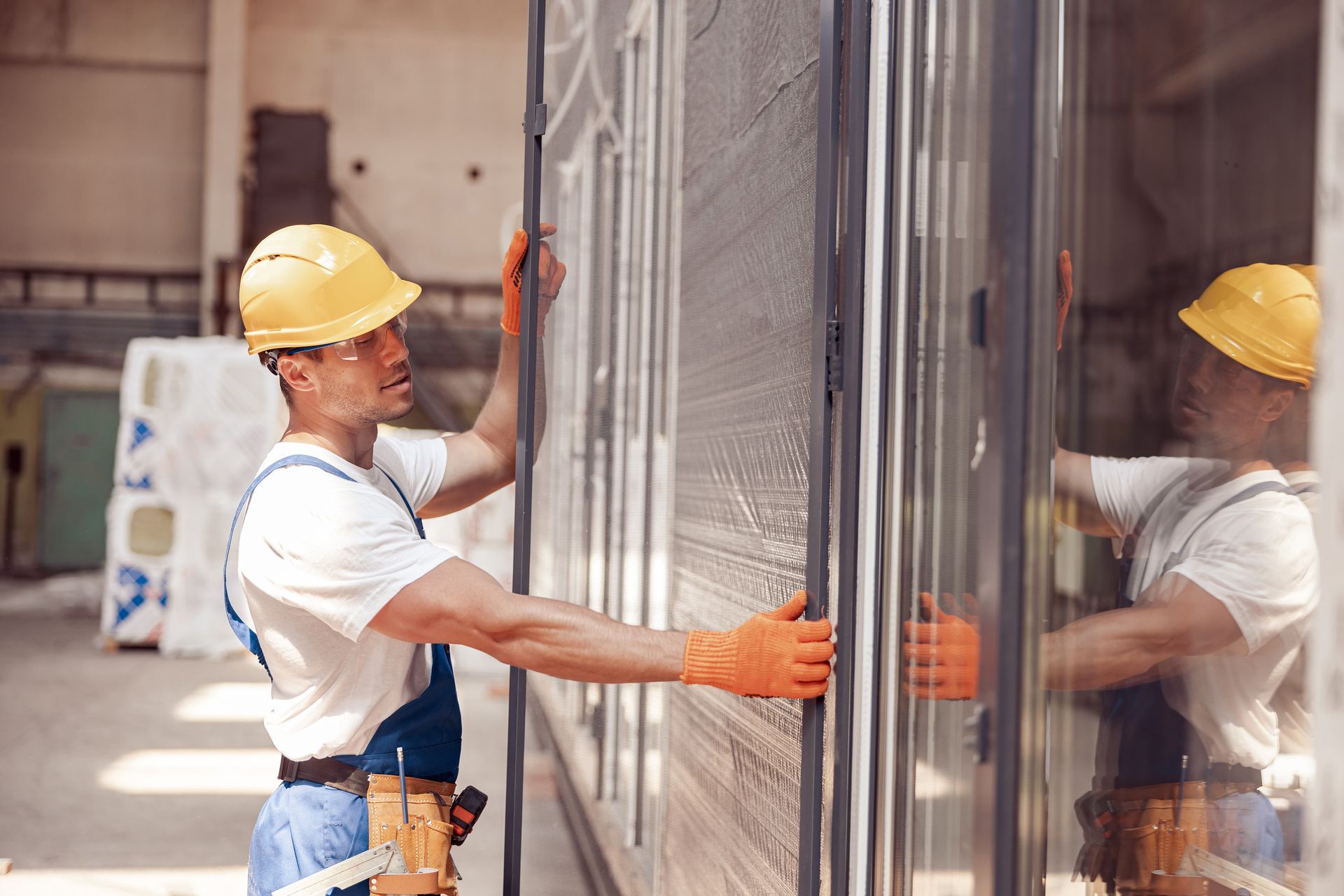 Worker in yellow hard hat and orange gloves installing a large glass panel on a construction site.
