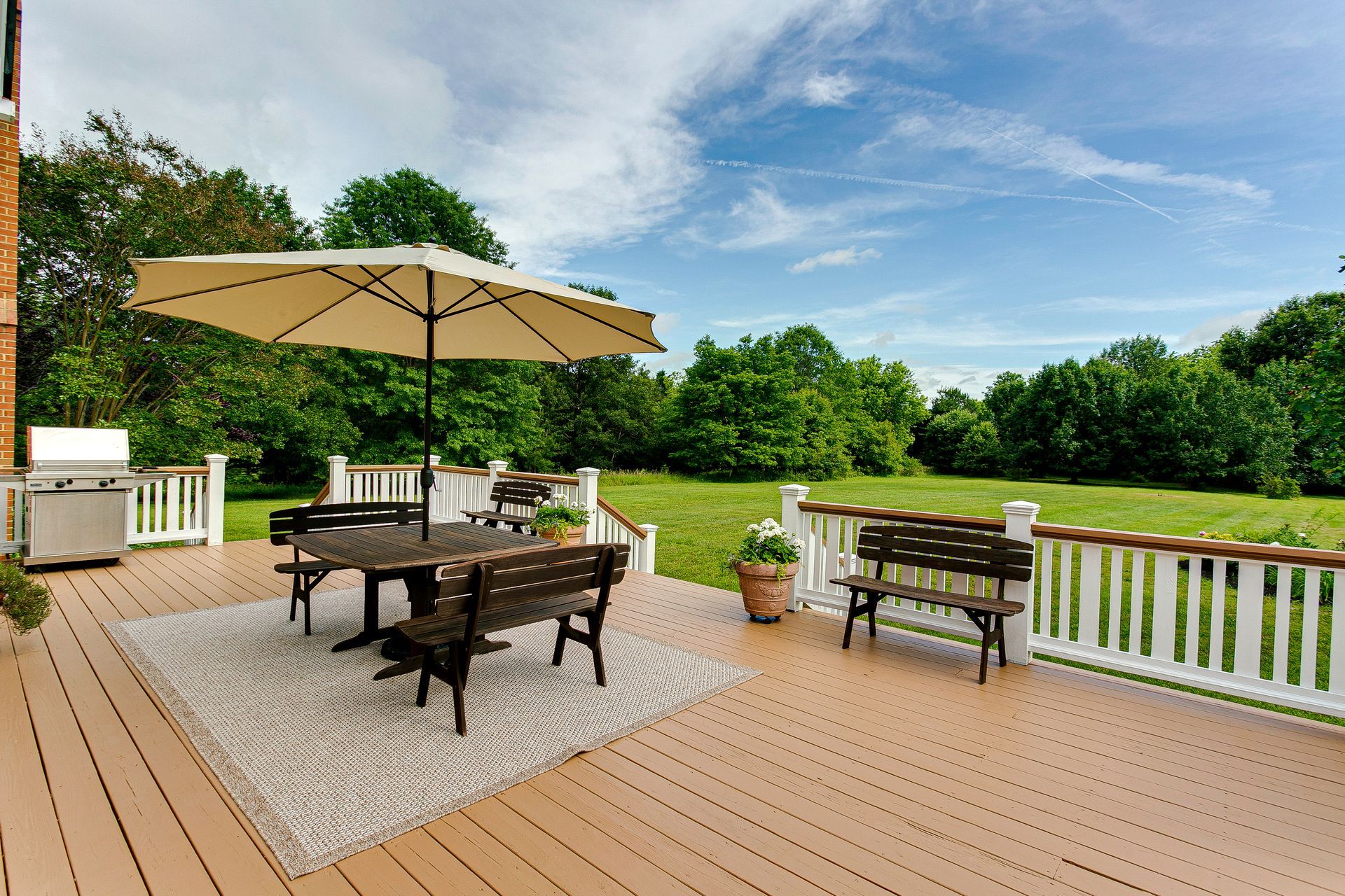 Backyard deck with dining table, umbrella, grill, potted plant, and a view of green trees and water.
