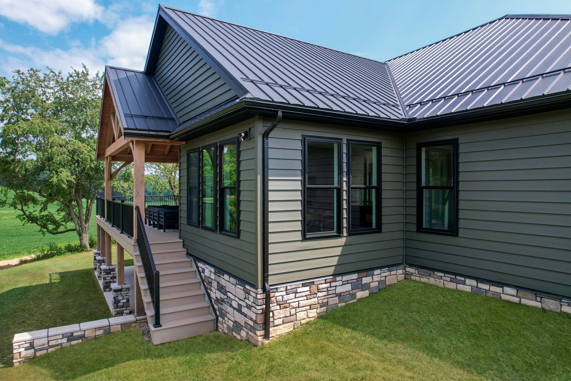 Modern house exterior with dark metal roof, stone base, and covered porch on a grassy lawn