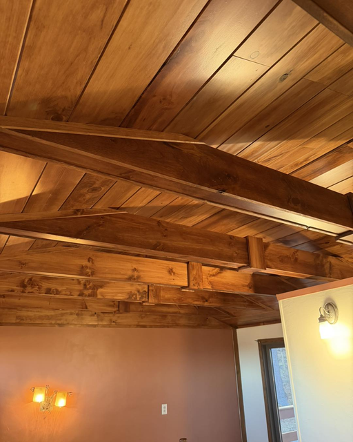 Wood-paneled ceiling with exposed wooden beams above a room with warm wall lights.