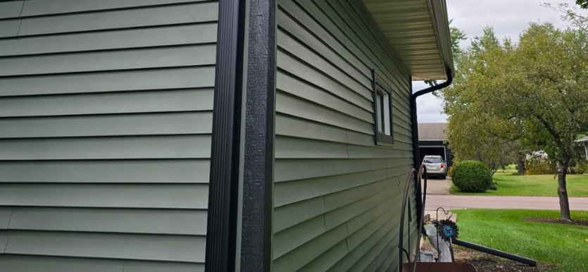 Side view of a green house with gray siding beside a lawn and trees on an overcast day