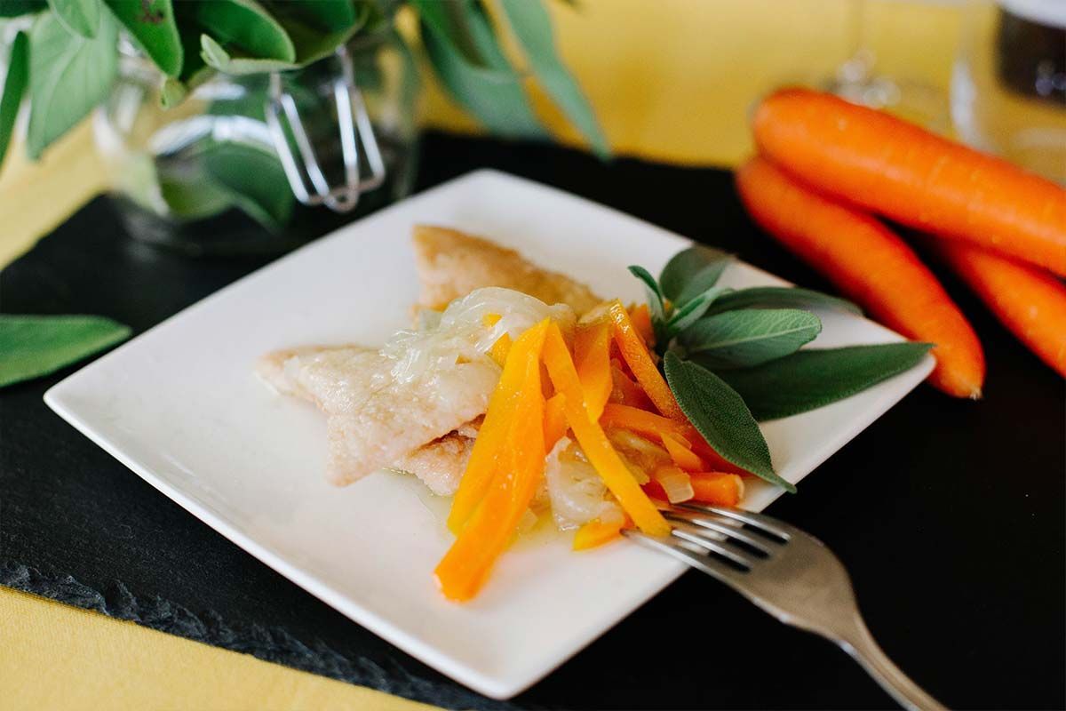 Pesce impiattato con carote a fette e verdure a foglia verde su un piatto quadrato bianco, con carote intere a lato.