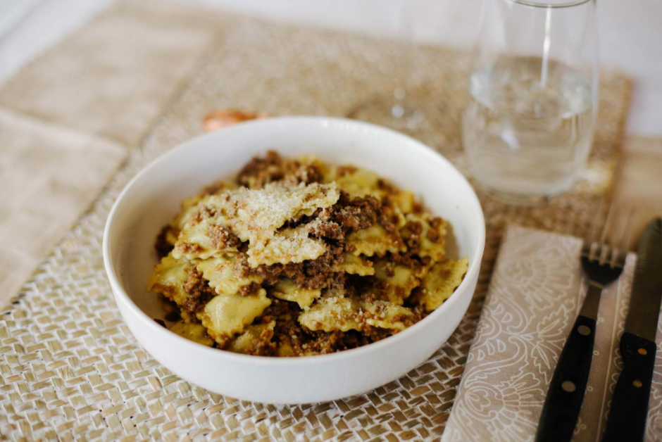 Una ciotola di pasta al forno con formaggio grattugiato su una tavola apparecchiata con forchetta, coltello e bicchiere.
