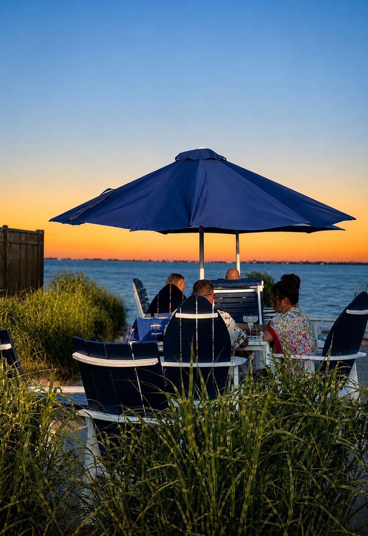 People dining under a blue umbrella by the beach at sunset