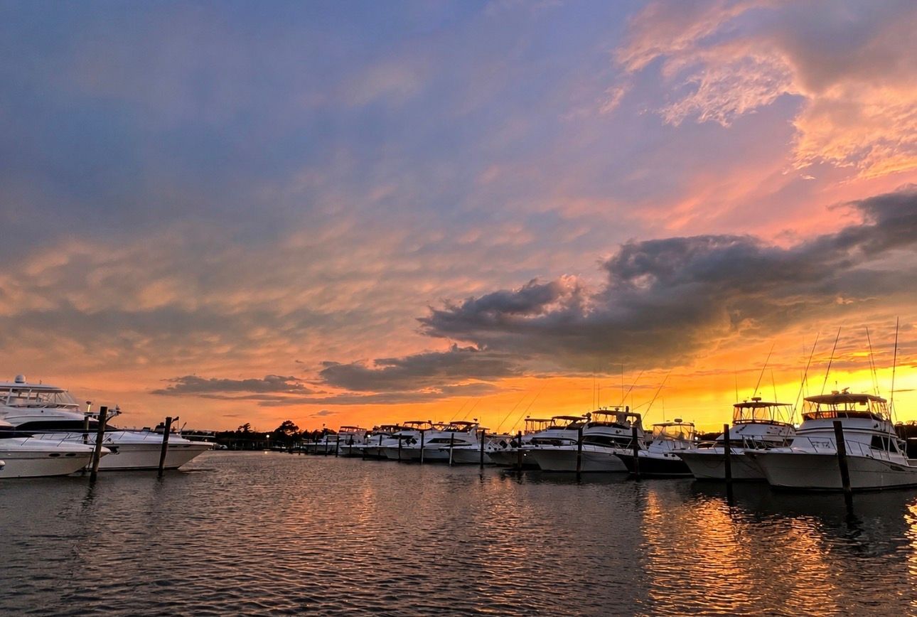 Boats docked on calm water at sunset, with orange and purple clouds glowing over the harbor.