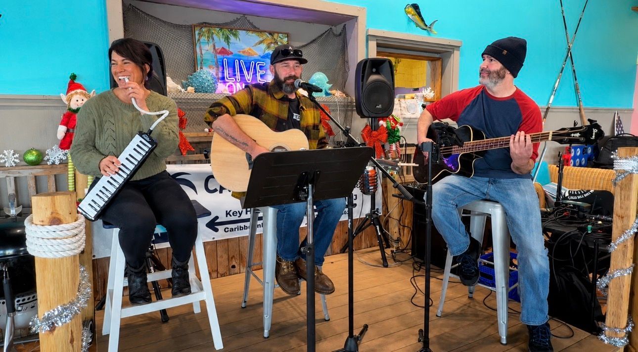 Four musicians perform indoors with guitars, keyboard, and drums in a colorful room.