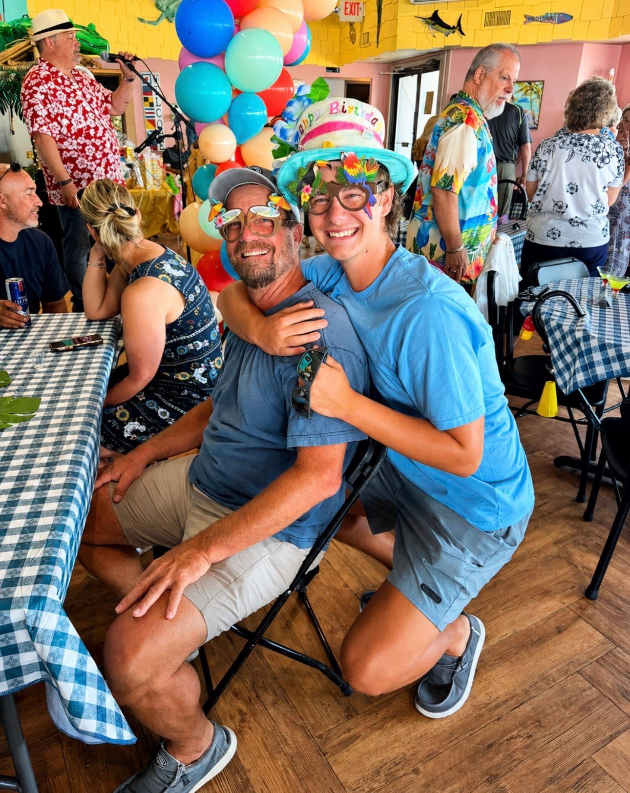 Two smiling people in colorful party hats pose at a festive indoor gathering with balloons and checkered tablecloths