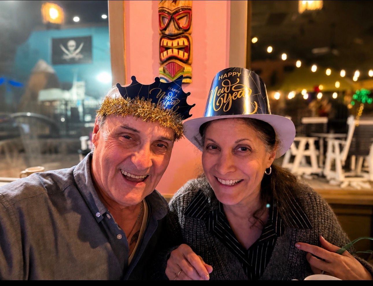 Two smiling people in festive hats pose in a warmly lit restaurant at night