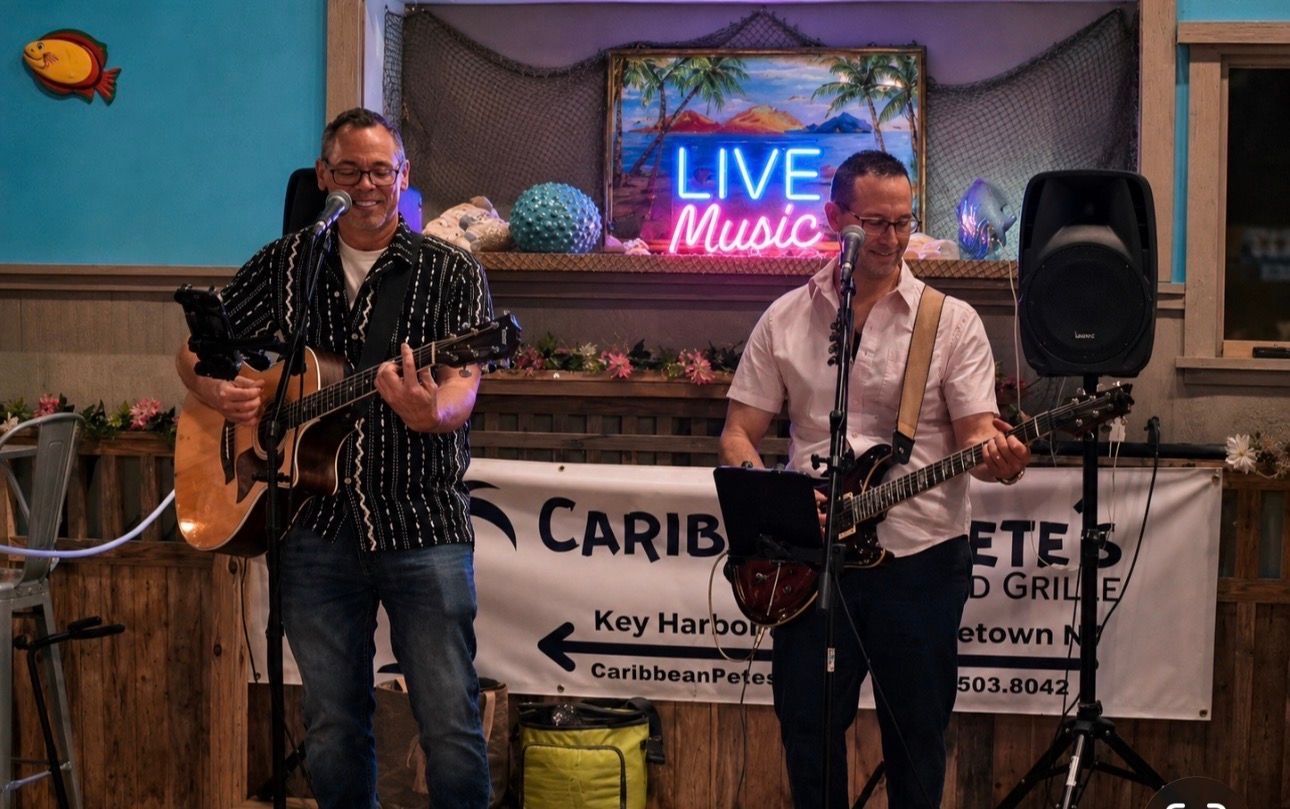 Two musicians perform live on a small stage with guitars and a neon “LIVE Music” sign.