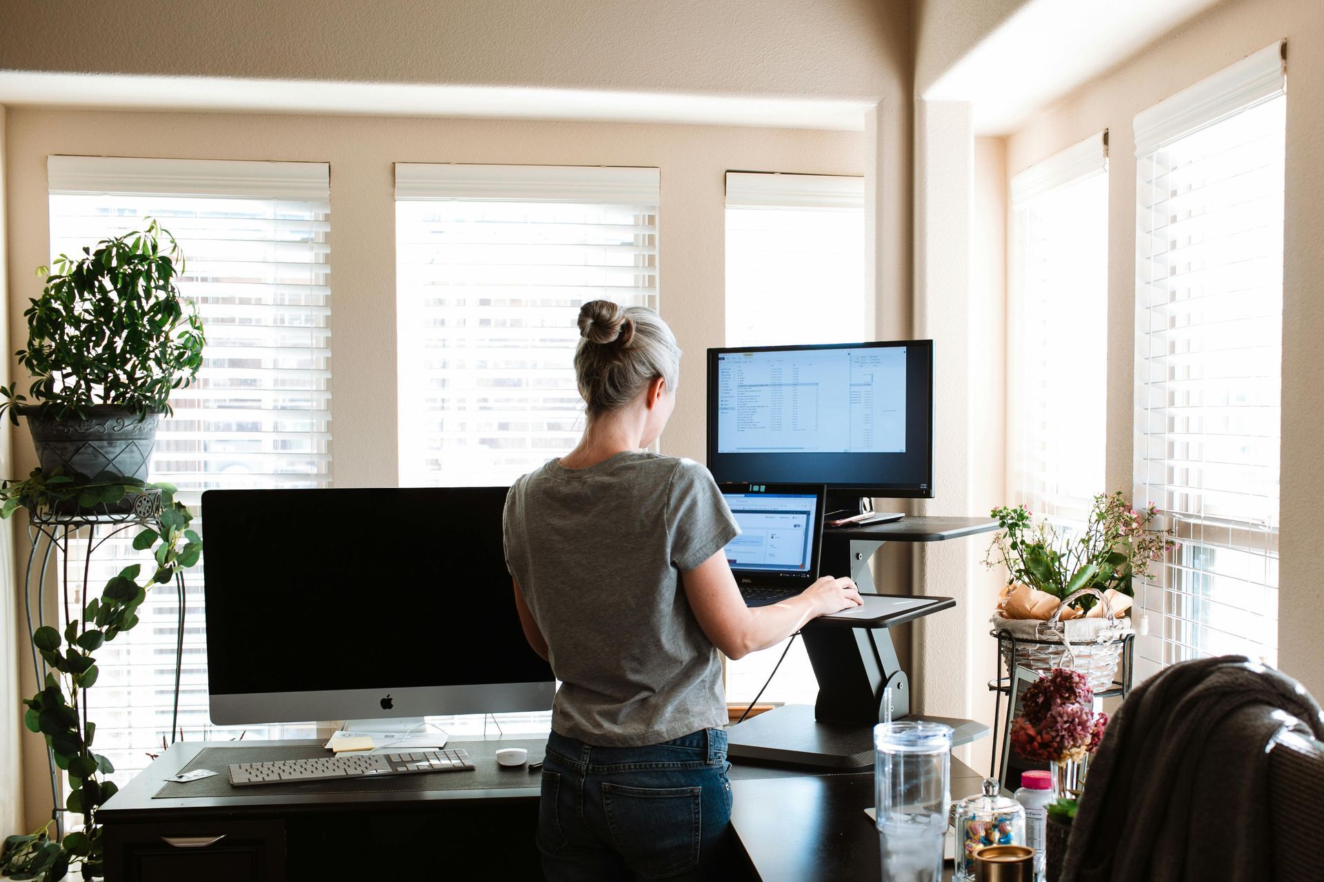 Woman standing at a standing desk, working on computer monitors in a bright room.