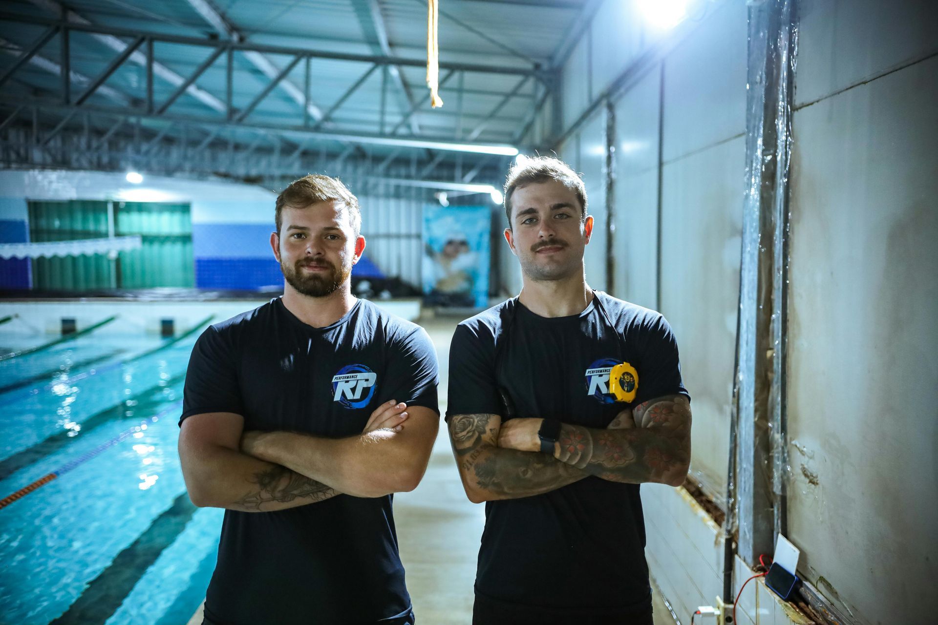 Two men stand in an indoor pool area. They wear black shirts and have tattoos, arms crossed. The pool is blue.