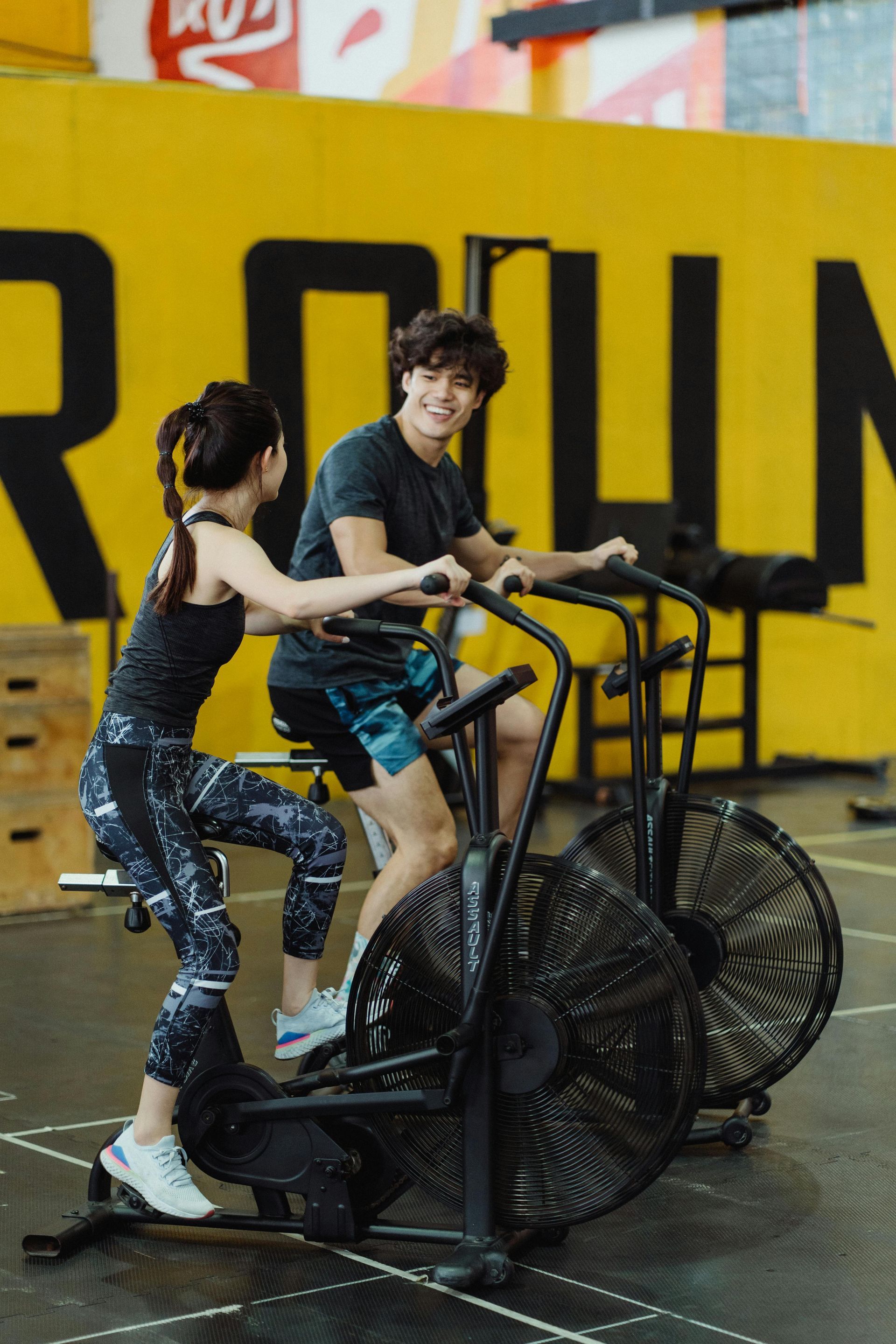 Two people on stationary exercise bikes in a gym. Man smiles, woman by his side. Yellow wall.