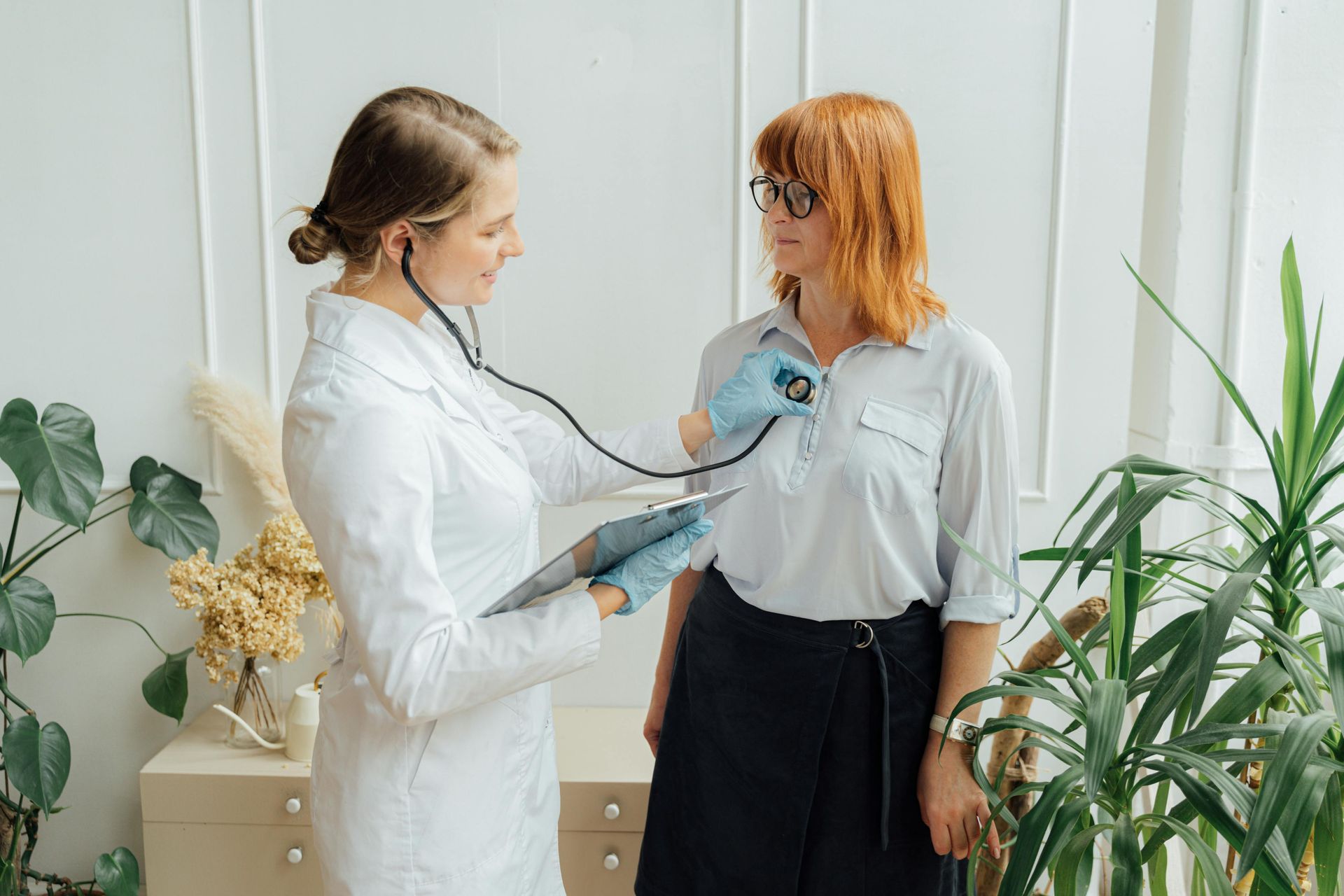 A doctor with a stethoscope examines a patient's chest in a light-filled room with plants. Both are looking at each other.