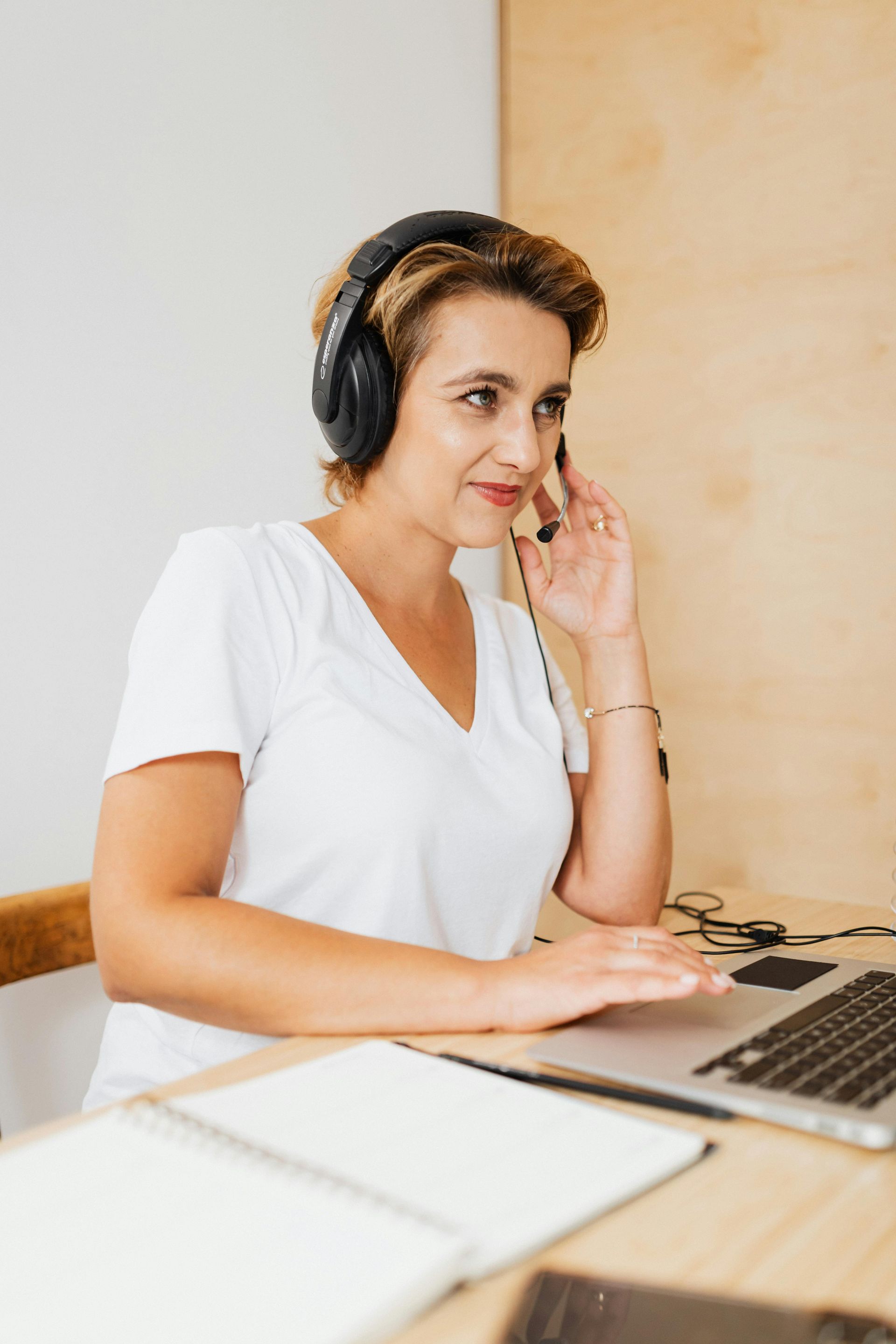 A woman wearing headphones is sitting at a desk talking on a cell phone.