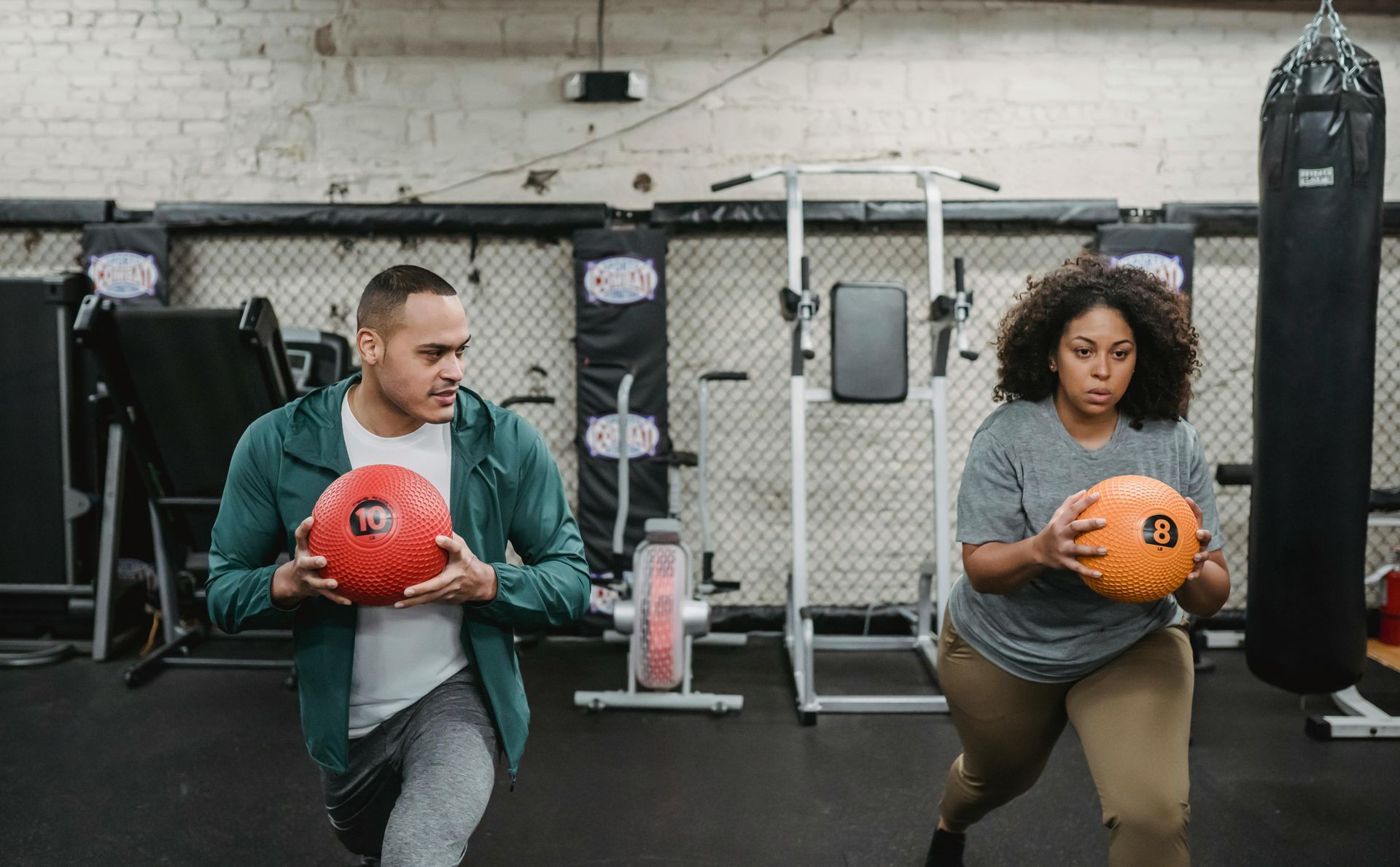 A man and woman are lunging in a gym, each holding a medicine ball. The man looks left; the woman focuses forward.