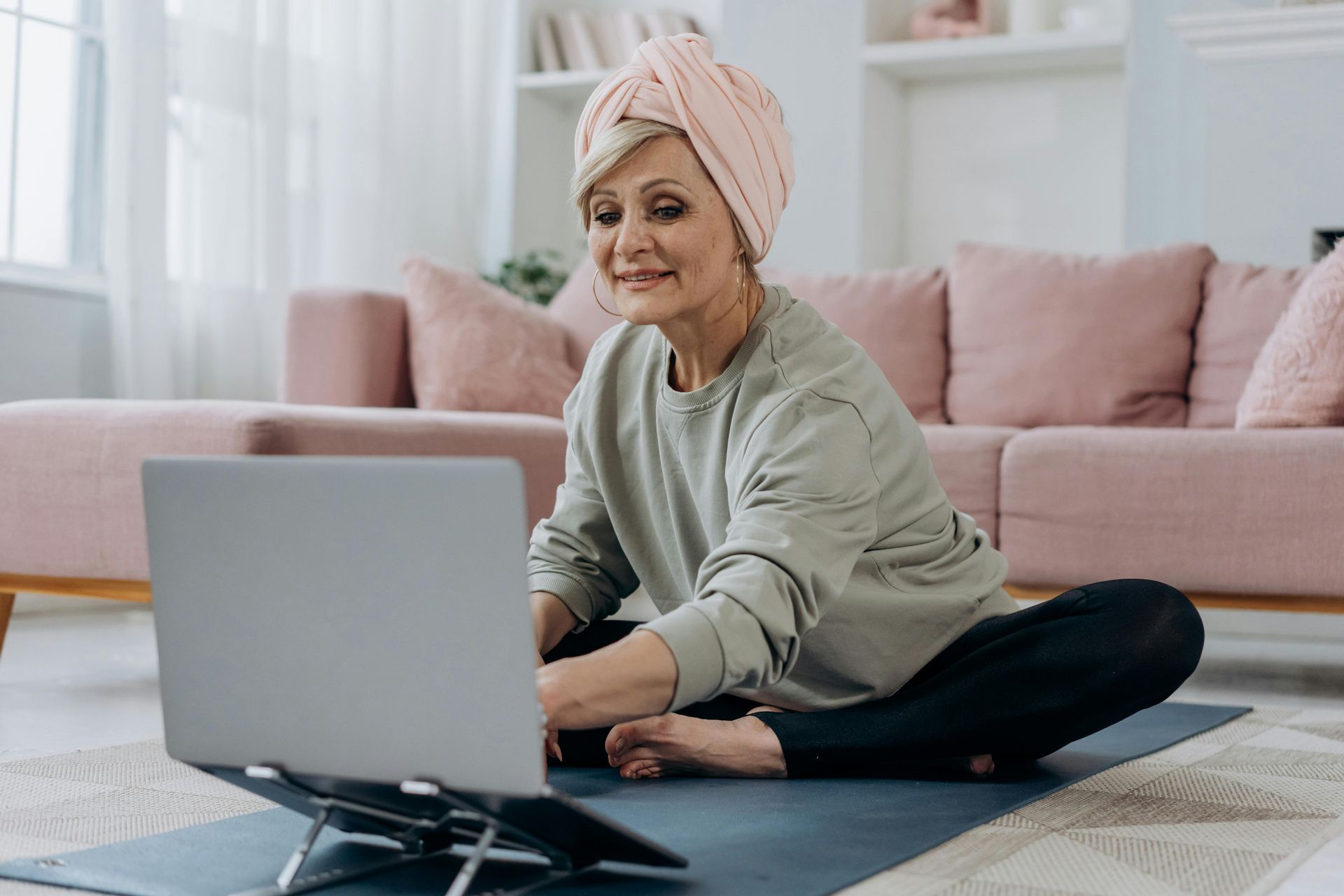 Woman in turban and activewear sits on yoga mat, using laptop in a living room, likely attending a virtual class.