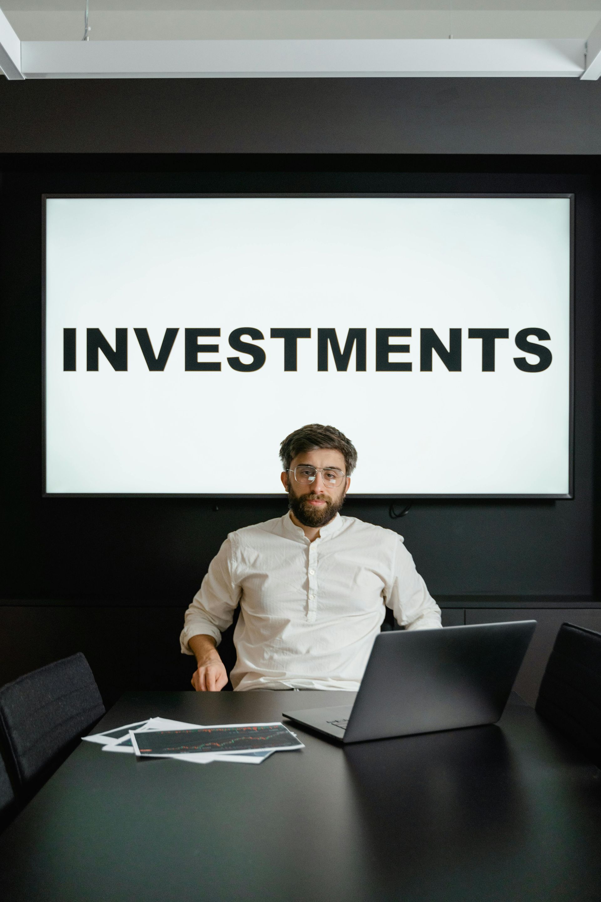 A man sits at a table with a laptop and papers in front of a screen displaying the word