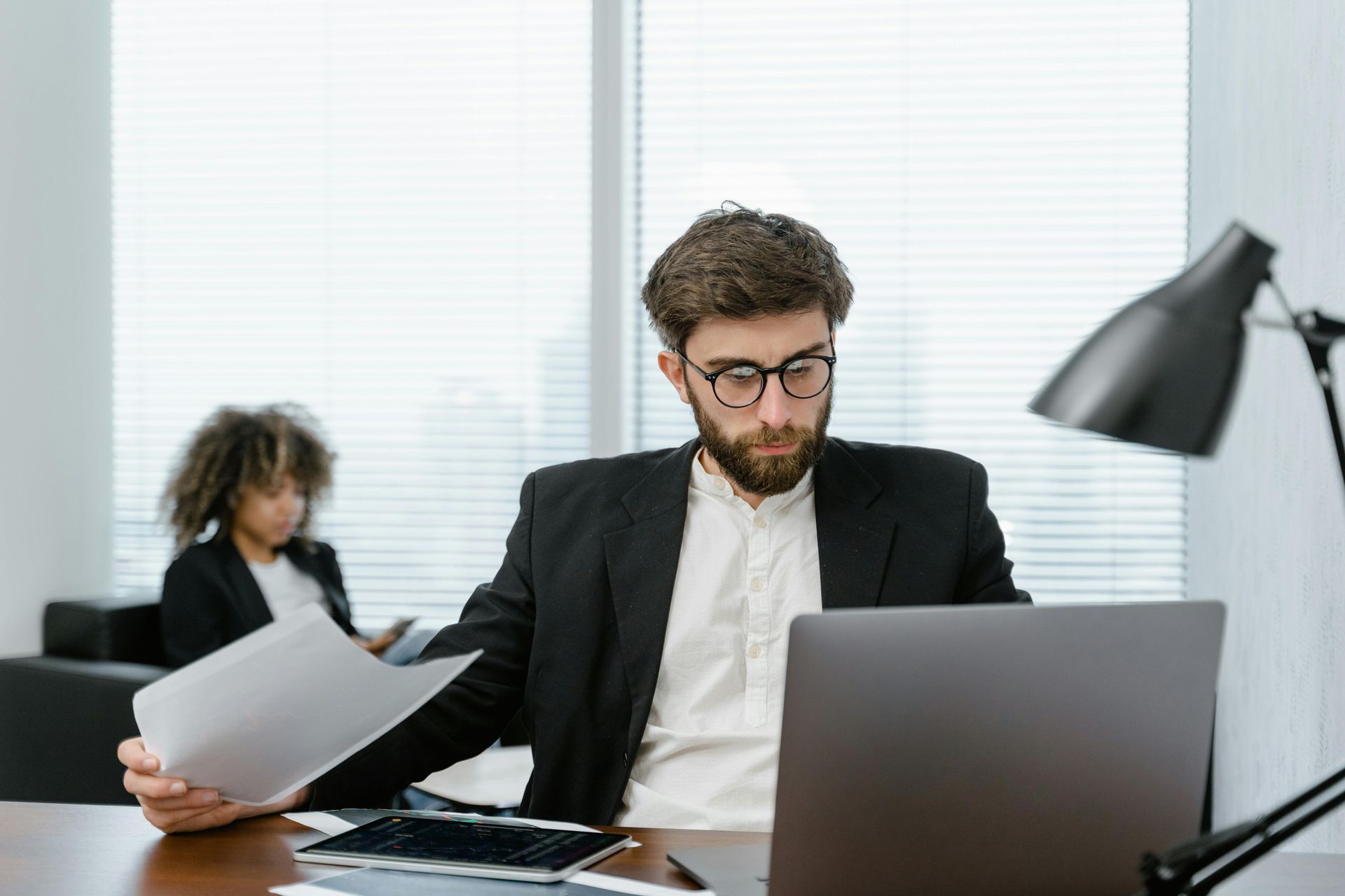 Man in glasses and suit examines papers at a desk with a laptop. Another person sits in the background.