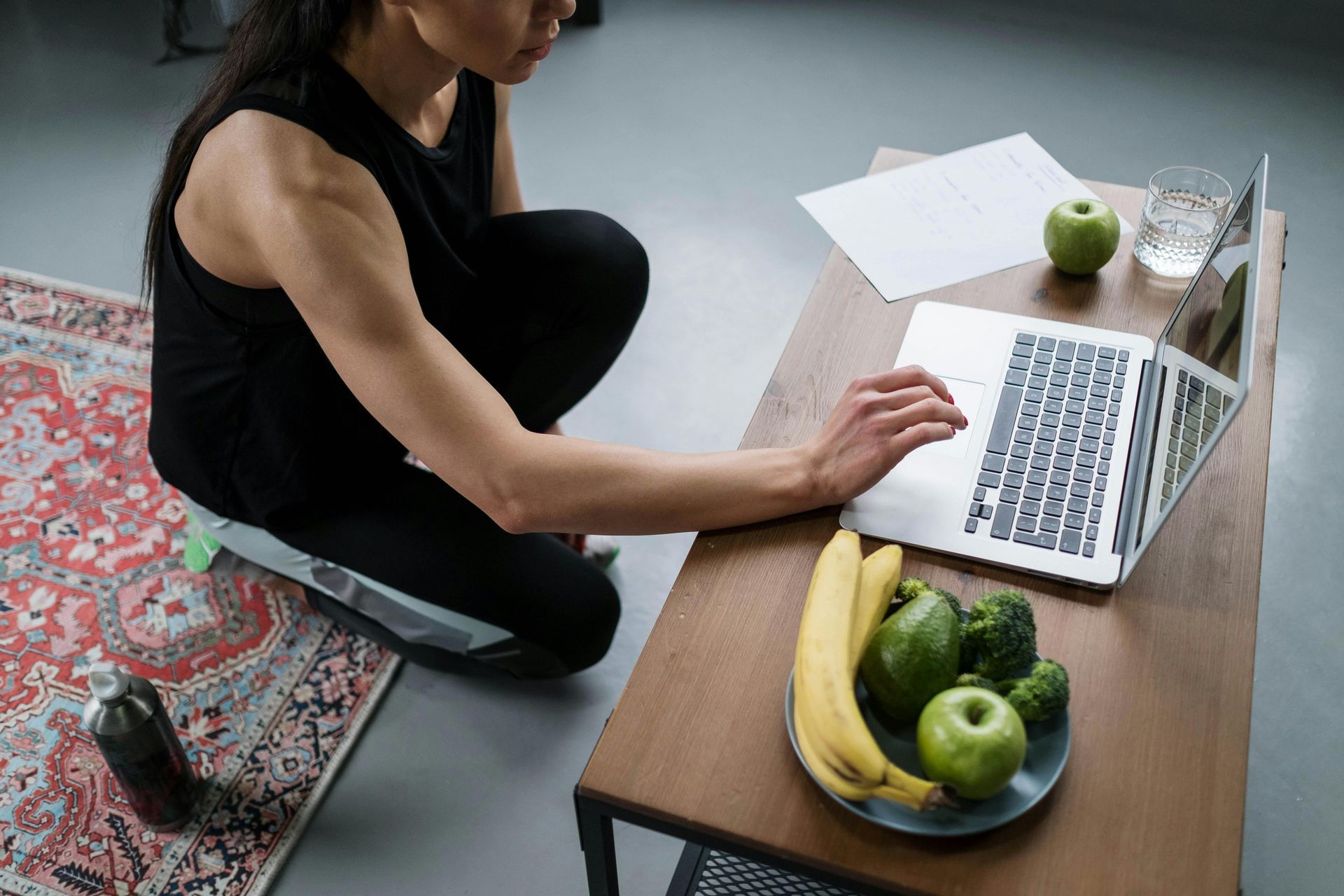 Woman working on a laptop while kneeling next to a table with a plate of fruit and a glass of water.