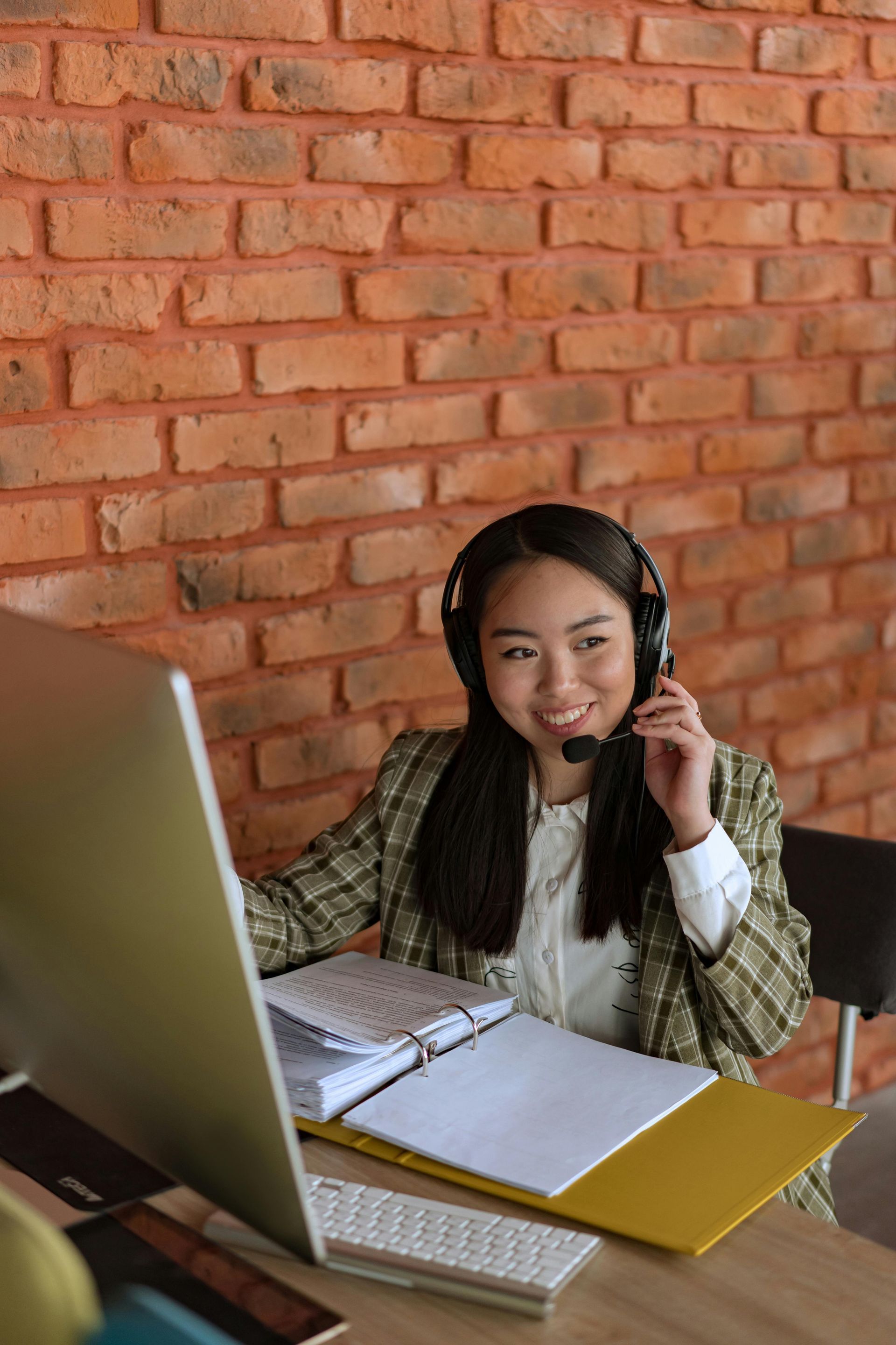 Woman with headset at desk, smiling. Brick wall background, computer, paperwork visible.