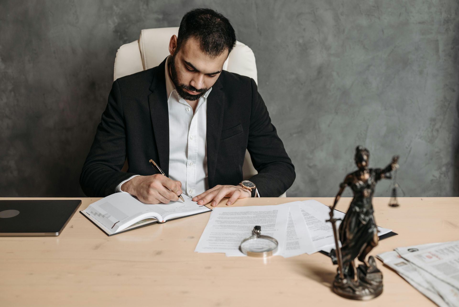 A man is sitting at a desk writing in a notebook next to a statue of justice.