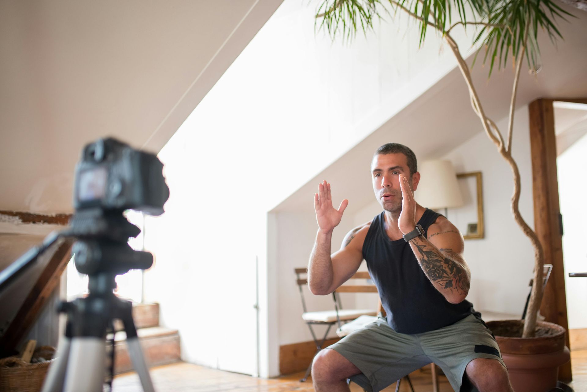 Man in workout gear, doing a squat, filming himself with a camera on a tripod indoors.