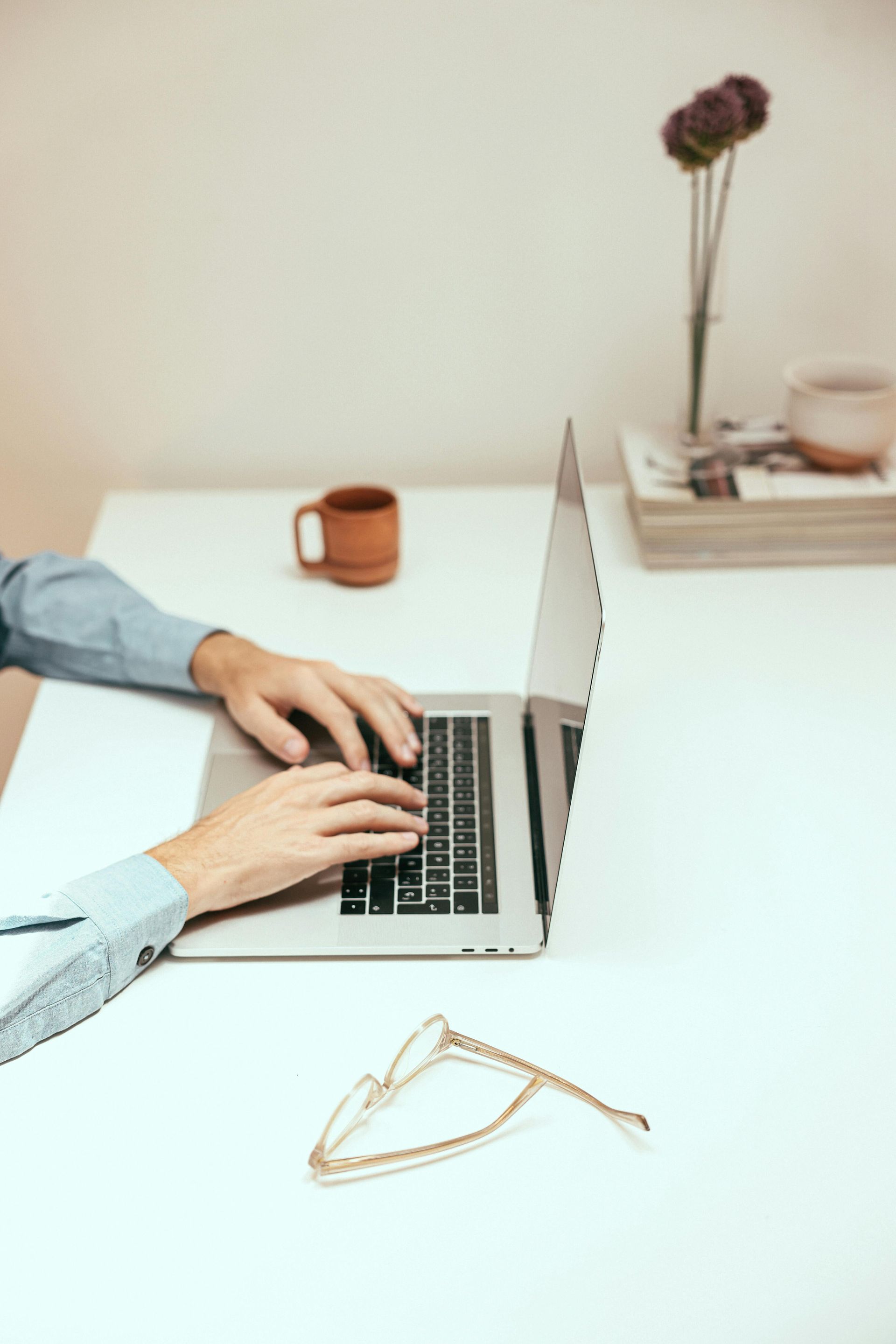 Person typing on a laptop at a white desk, with a mug, flowers, and glasses nearby.