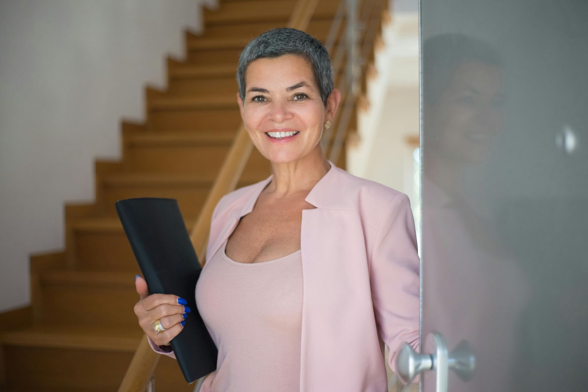 Woman with short gray hair, wearing pink blazer, smiles while holding a folder in a doorway. Staircase in the background.