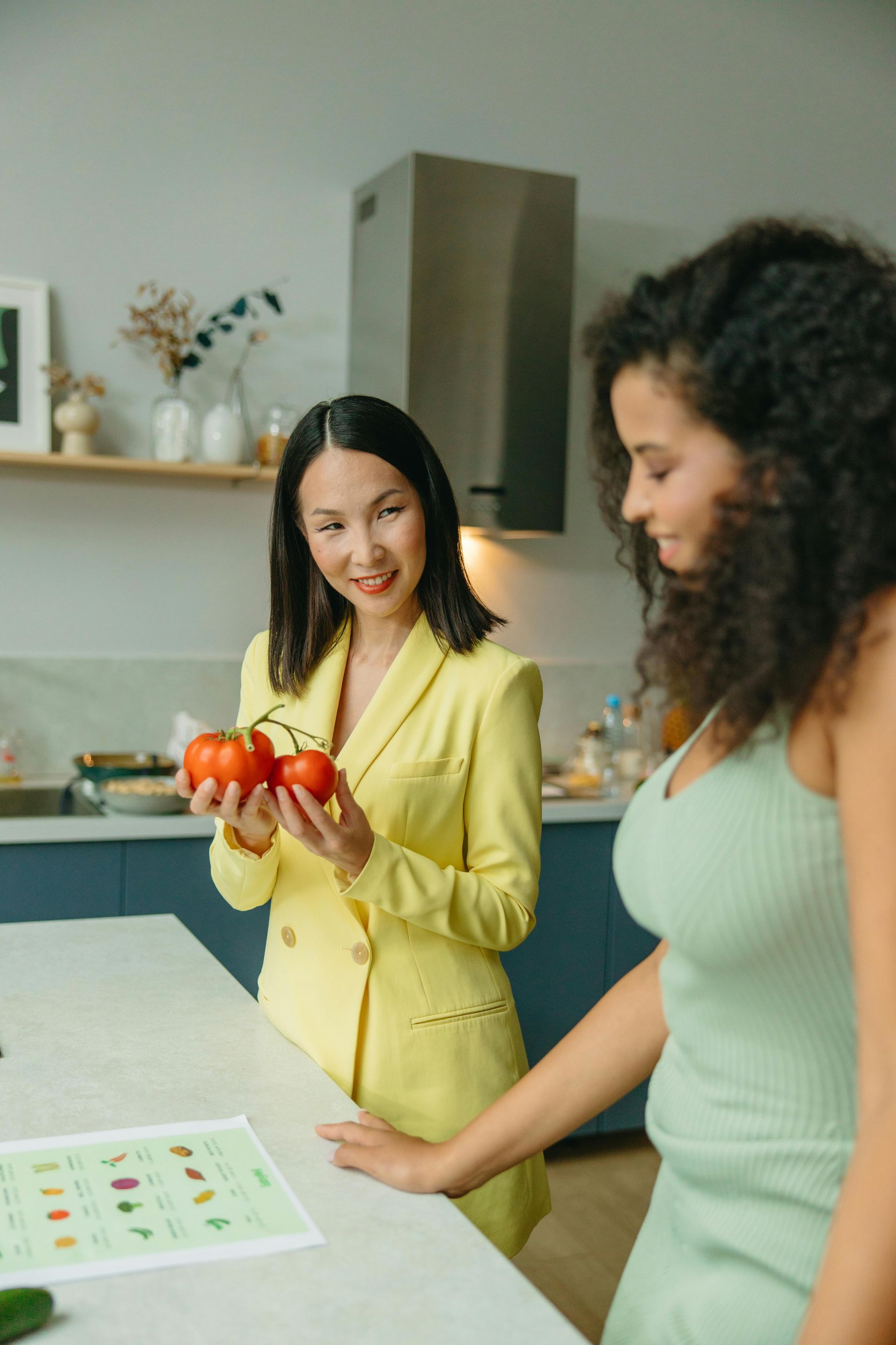 Two women in a kitchen, one holding tomatoes, smiling. The other looks down. They stand by a counter with a produce chart.