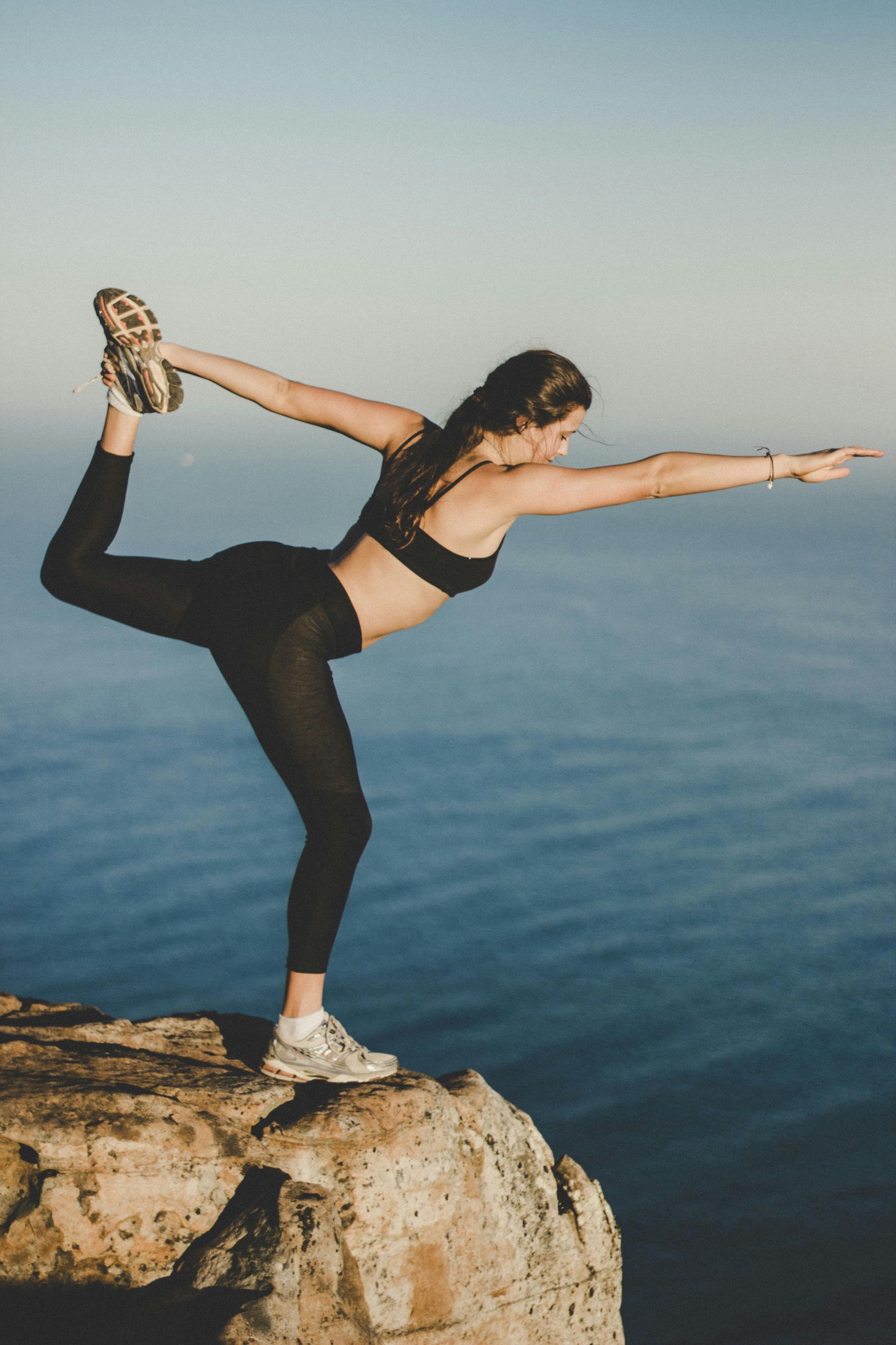 Woman in yoga pose on a cliff edge, balancing on one foot with a hand outstretched. Blue ocean and sky background.