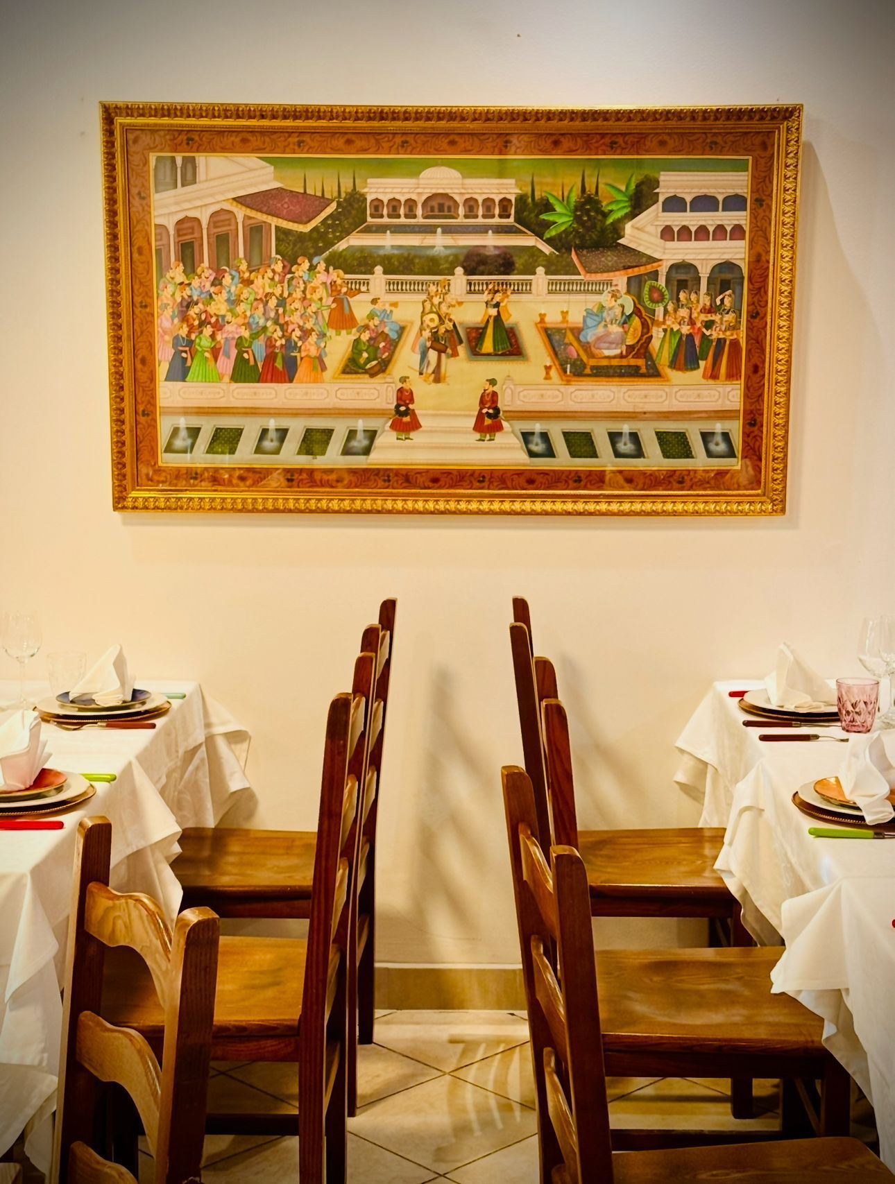Piccola sala da pranzo con due file di sedie di legno, tovaglie bianche e un arazzo colorato alla parete.