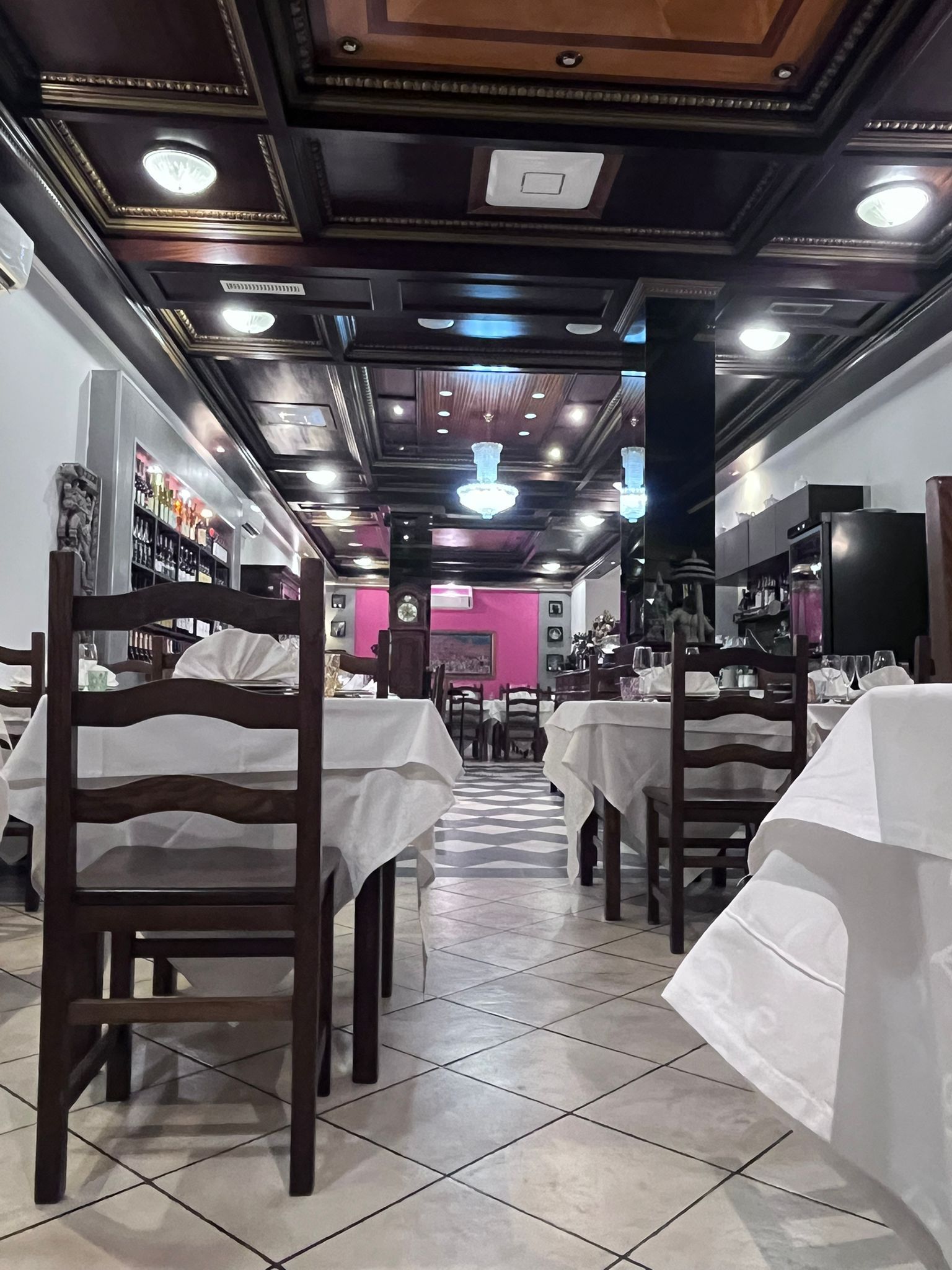 Interno vuoto di un ristorante con tovaglie bianche, sedie scure, pavimento piastrellato con motivi decorativi e lampadari decorati sul soffitto.