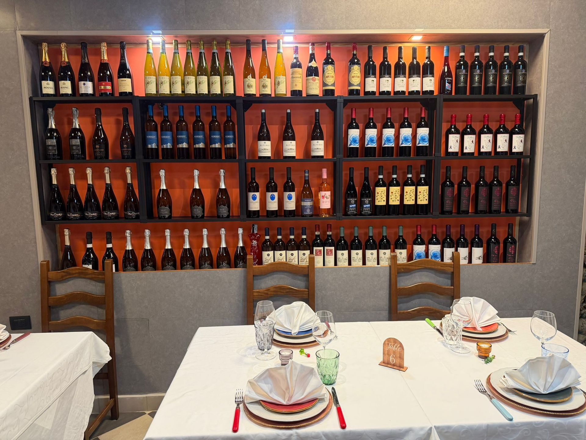 Scaffali di bottiglie di vino nella sala da pranzo di un ristorante con tavoli apparecchiati e tovaglioli piegati.