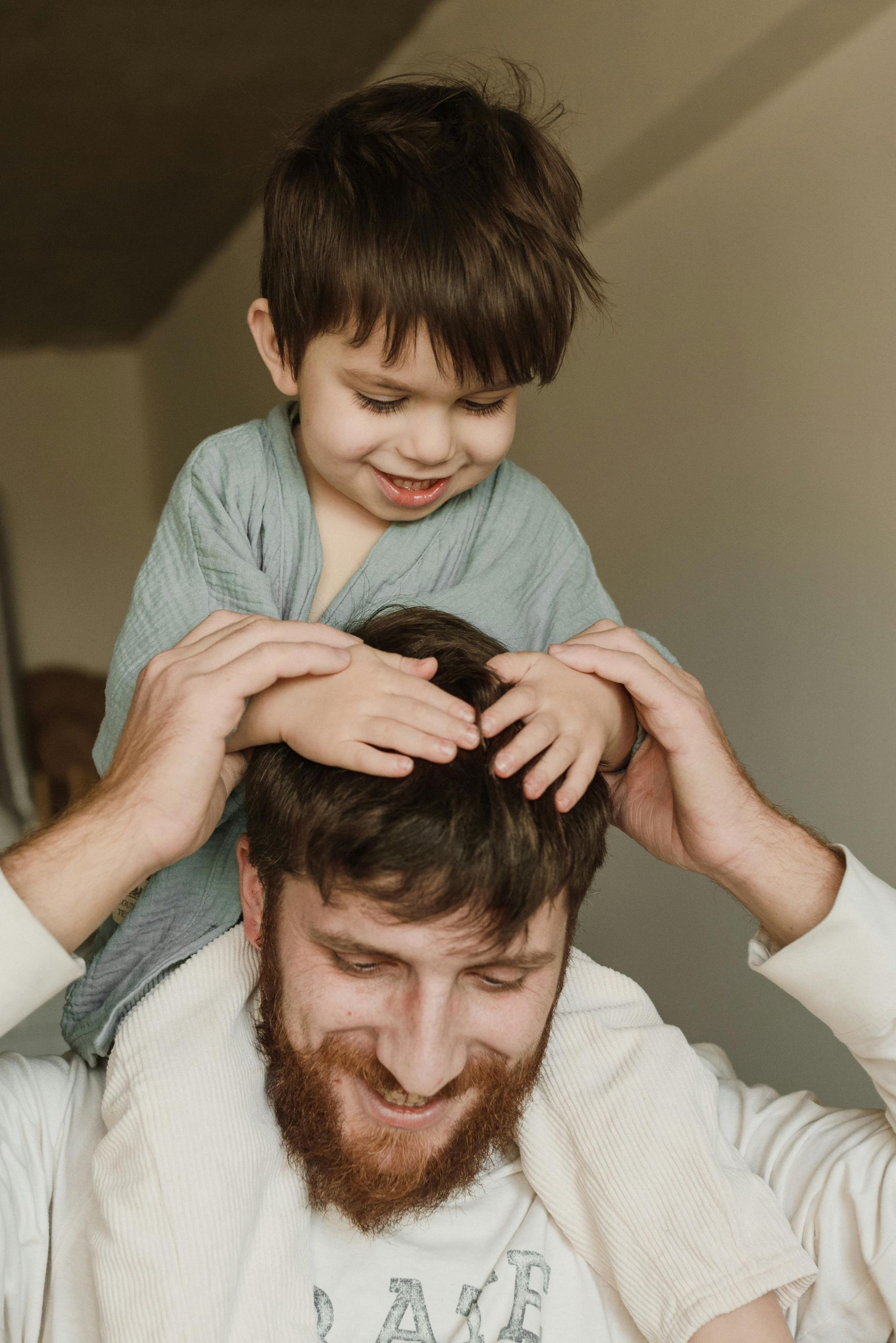 A young child, smiling, sits on a man's shoulders, resting hands on his head. Both are indoors.