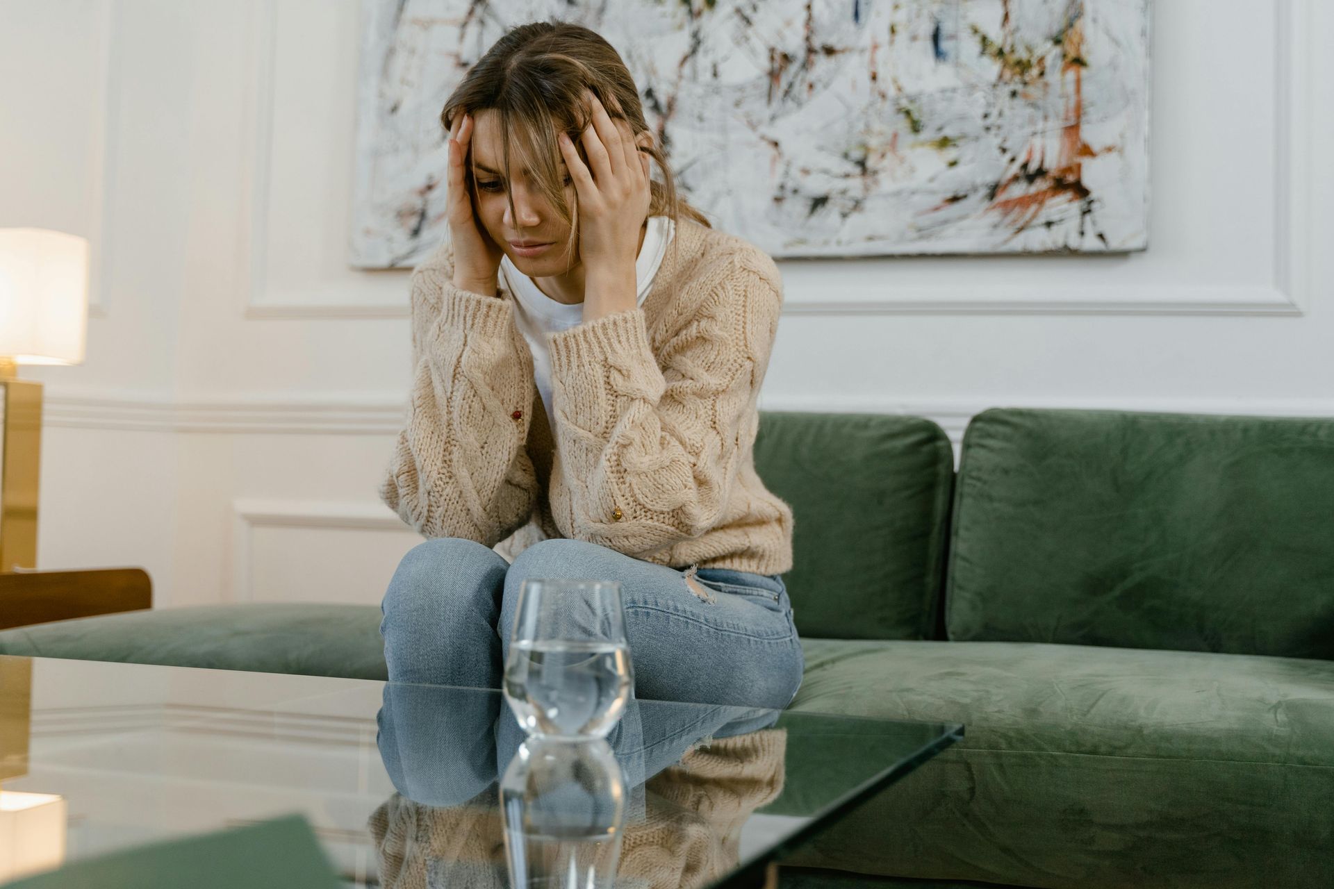 Woman with hands on her head, looking down, sitting on a green couch with a glass of water in front of her on a coffee table.